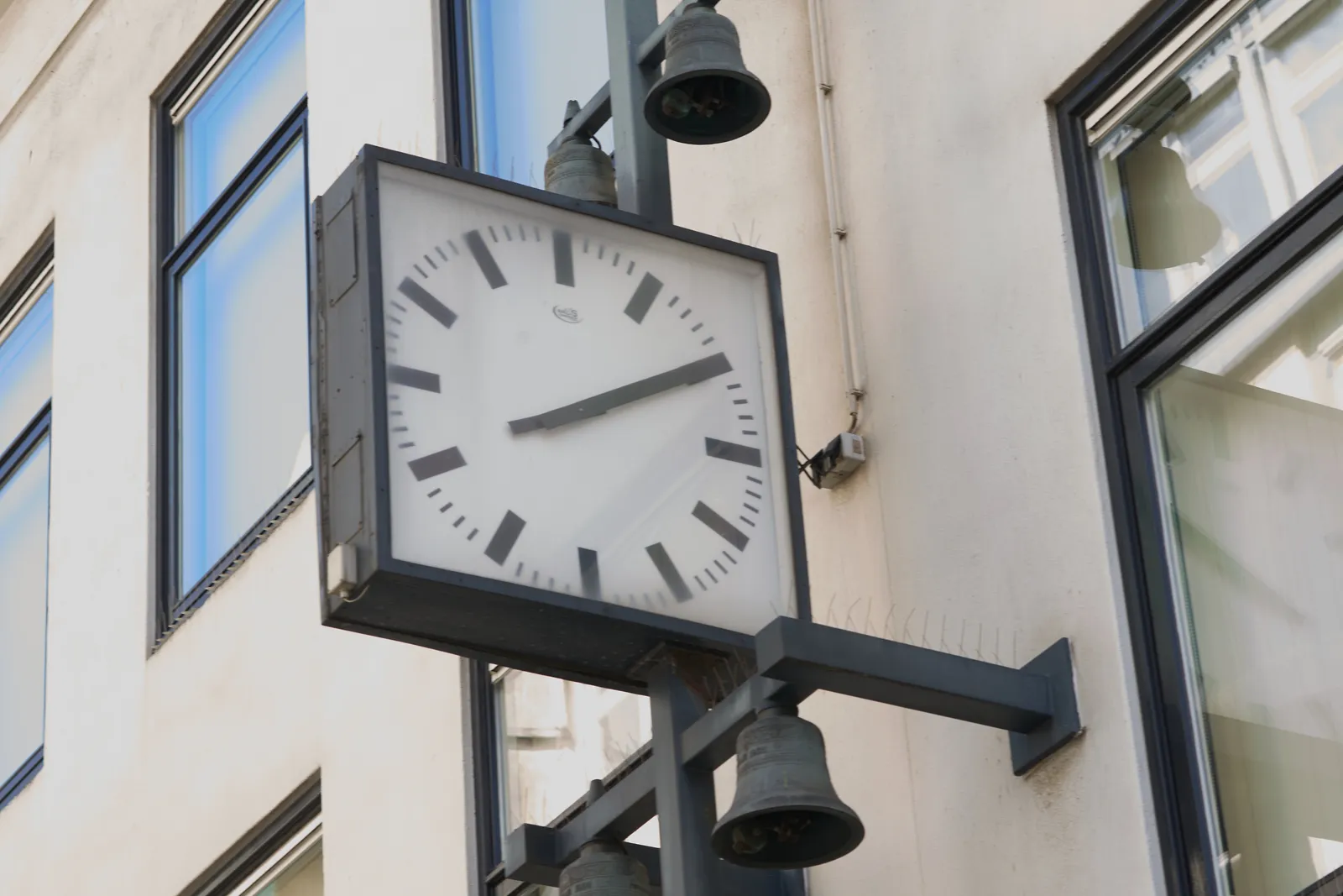 A black and white building, with attached to it a modern sculpture. It’s made from strong vertical lines, with bells, and a square clock. This clock points at 10 past 8.