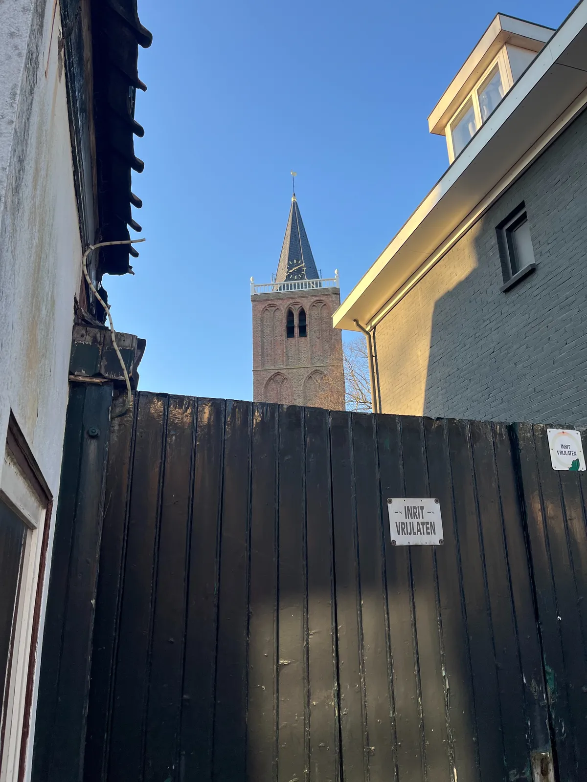 Peeking over a fence, between two small Dutch town houses, at a church tower. The clock on this tower points at 10 past 8.