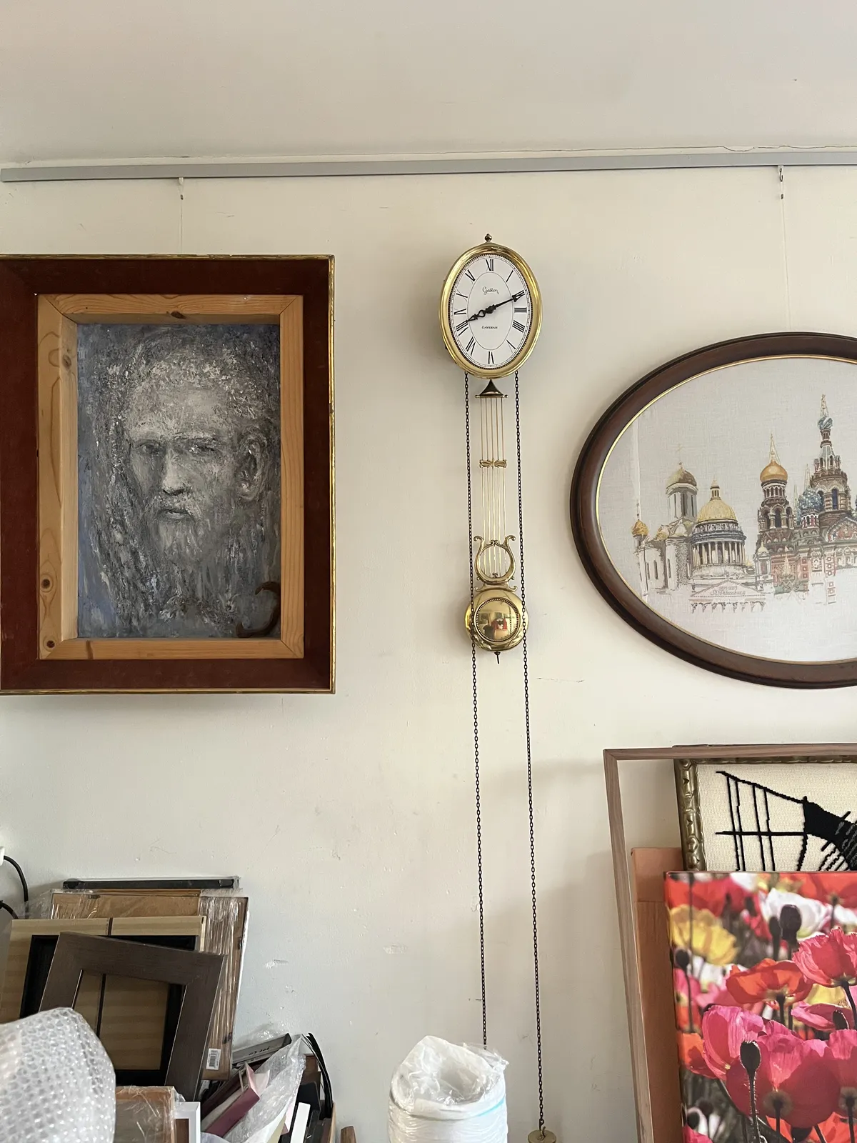 Between some paintings, in an unorganised space, there’s an old, golden wall clock with weights hanging from very long chains. It points at 11 minutes past eight.