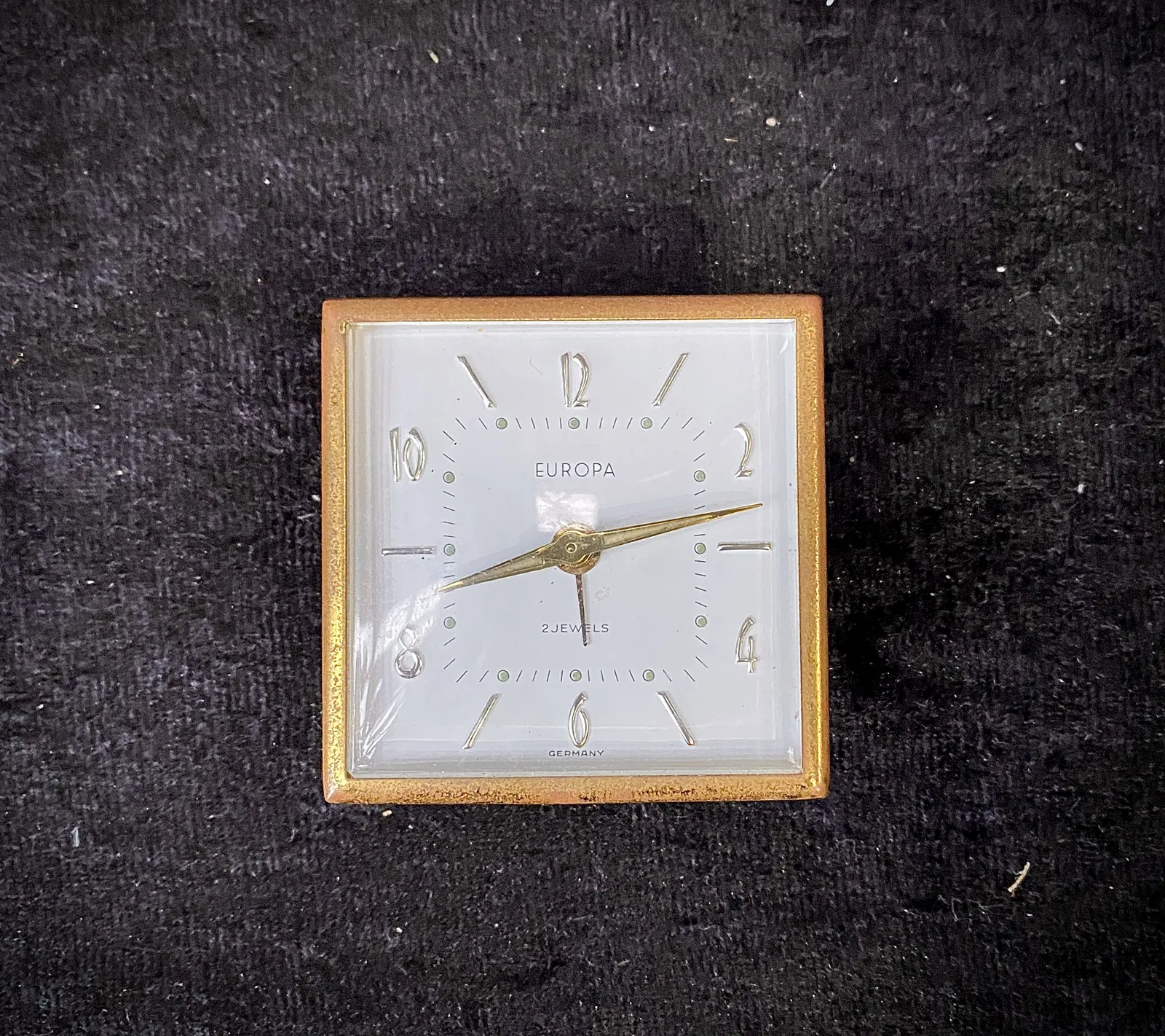 A square clock with the words Europa 2 jewels Germany printed on its face. It points at 13 minutes past 8.