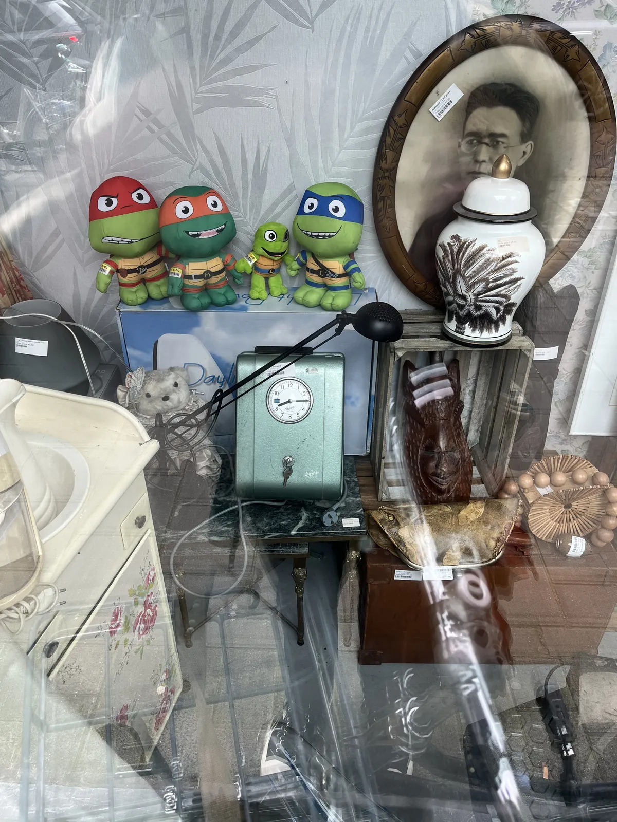 The shopping window of a thrift store, full of stuff. In the middle there’s an old mechanical, industrial looking clock which points at 14 minutes past eight.
