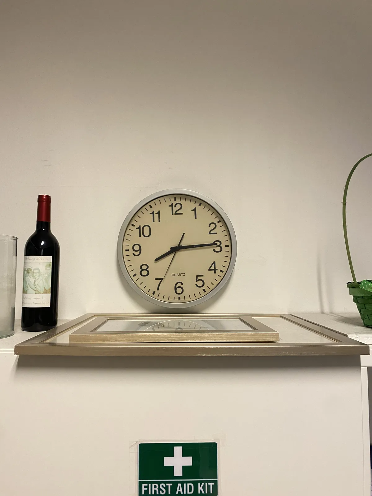 Two frames on an edge, you can’t see what picture is in it. There’s also a bottle of wine and a green sign that says First Aid Kit. And right in the middle of this picture there’s a clock which points at 14 minutes past 8.