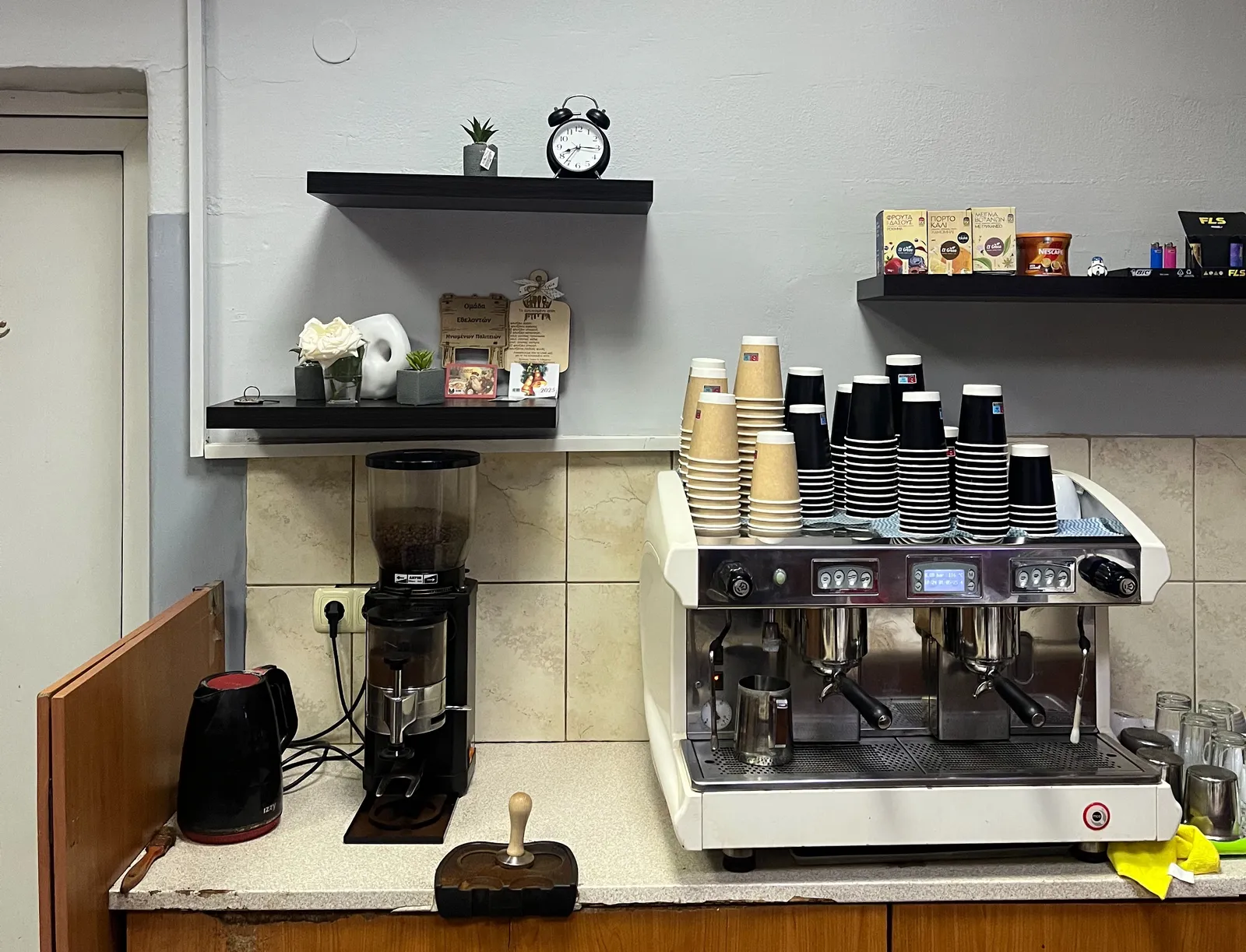 A professional coffee machine and a coffee grinder with some shelves over them. On one of these shelves there’s an old alarm clock that points at a quarter past eight.