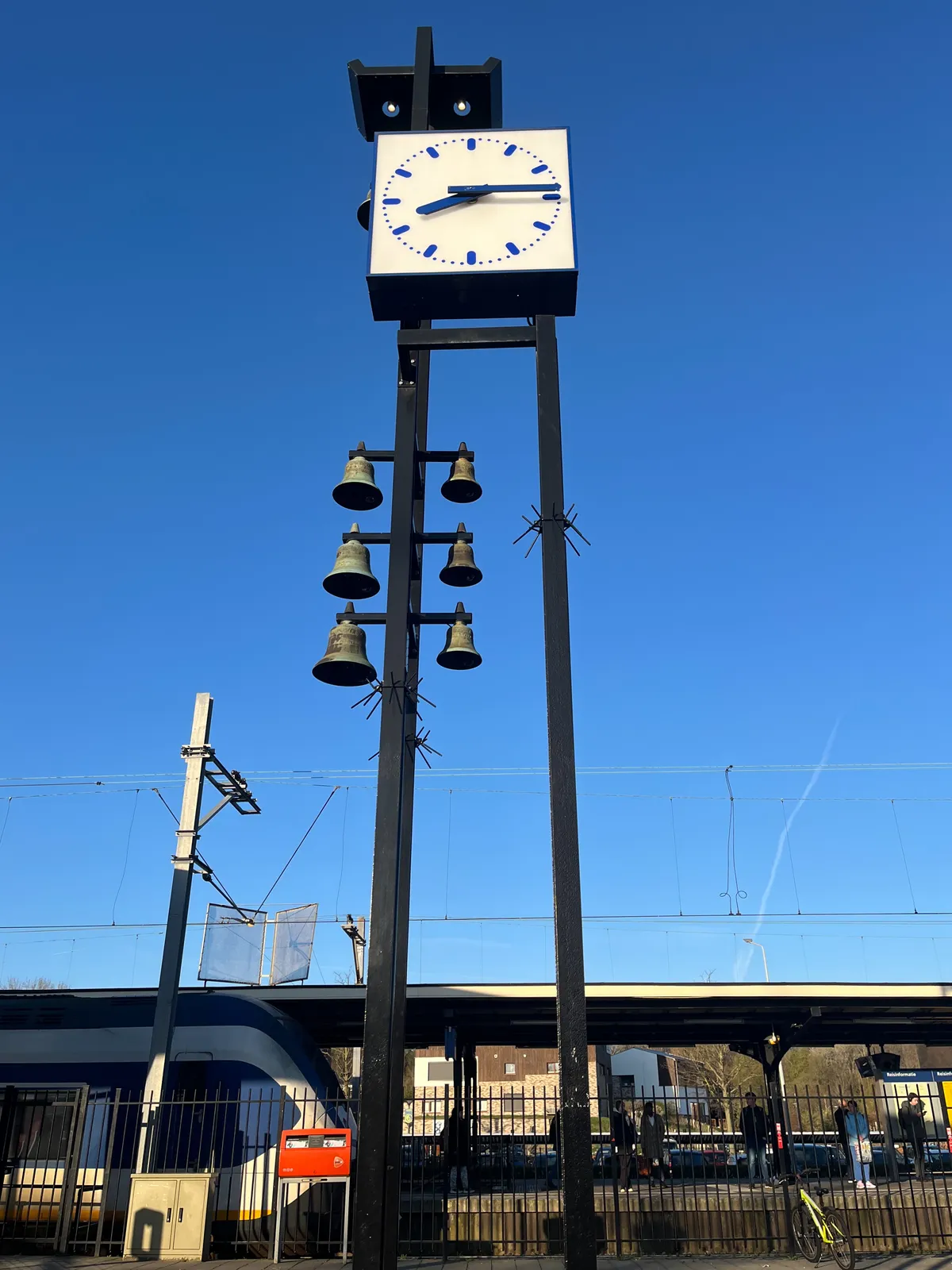 A Dutch station clock high on some black metal poles, against a bright blue sky. Attached to these poles are some bells as well. Behind it, at the bottom of the image, there’s a train. The clock points at a quarter past 8