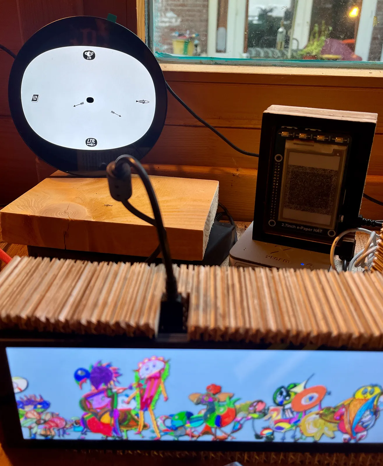 There are three digital devices in this picture. One of them, a rather wide screen, in the foreground, shows a bright parade of colourful illustrations. There’s a smaller screen in the background that shows an abstract black and white illustration. And then there’s a round screen that shows a rather minimalist clock with a tea pot, a fish, a book, and the letters LTR to indicate 12, 3, 6 and 9 o’clock. Its hands are smaller arrows. It points at 19 minutes past 8.