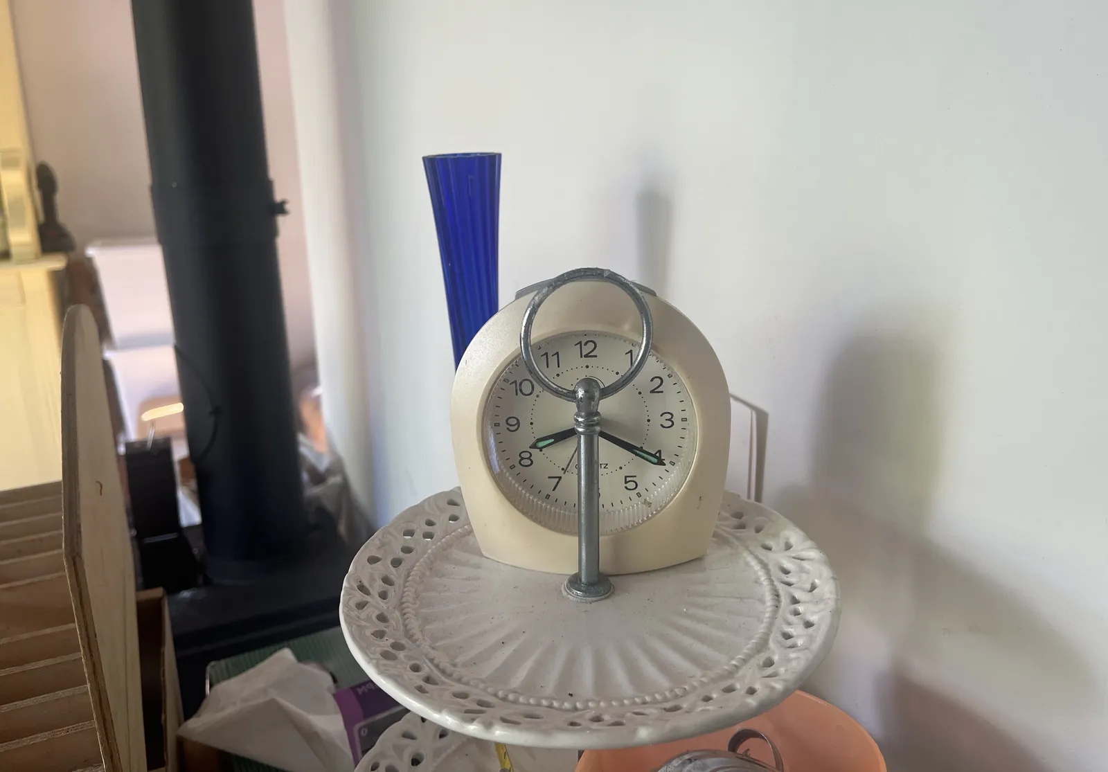 A little white alarm clock stands on top of a multilayered tray, behind the piece of metal for holding it. The piece of metal covers the center of this clock, but both hands clearly point out, to both sides, at 20 minutes past eight.