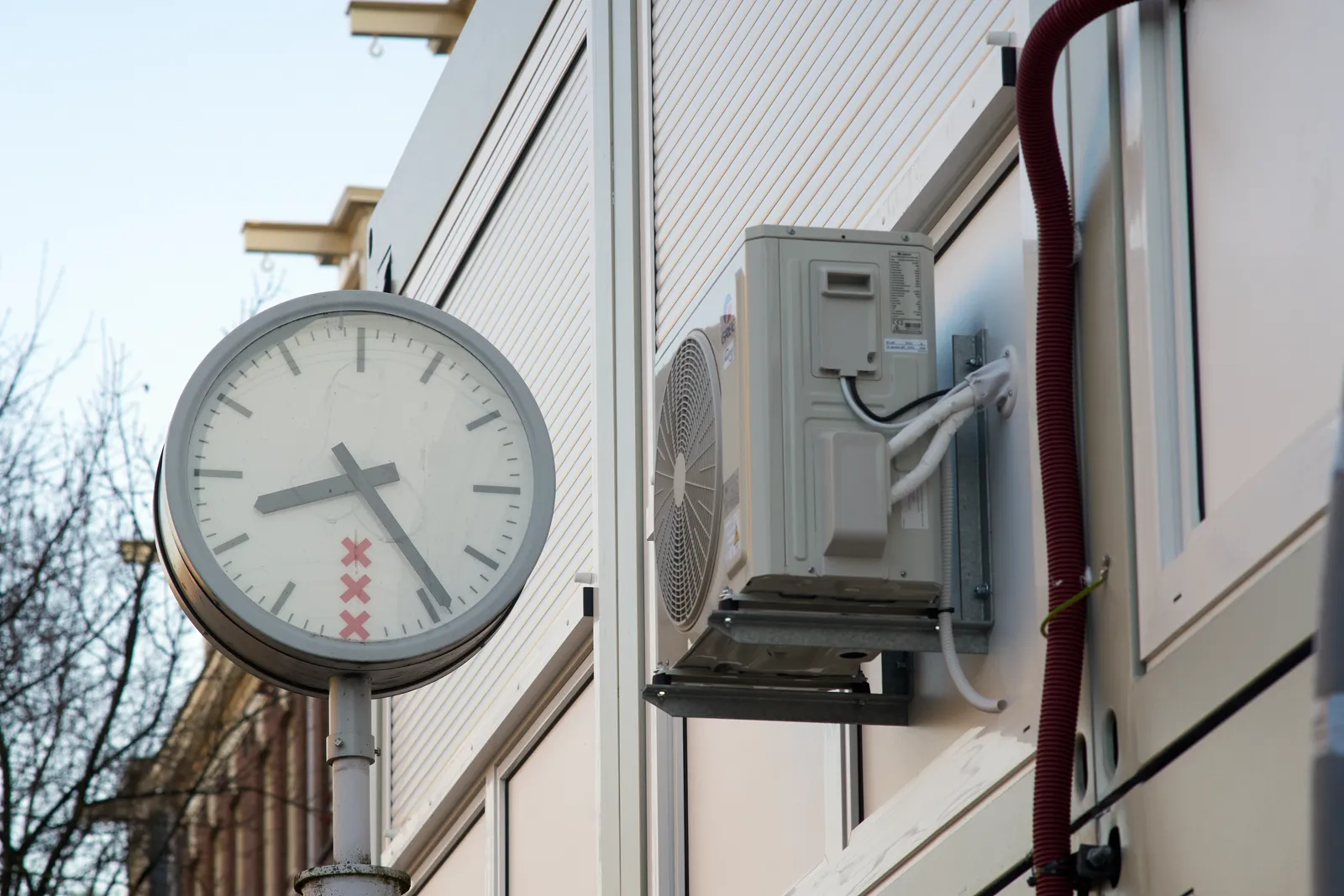 A municipal clock on a pole next to the external part of an airco, attached to a container unit. The clock on the pole points at 24 minutes past eight.