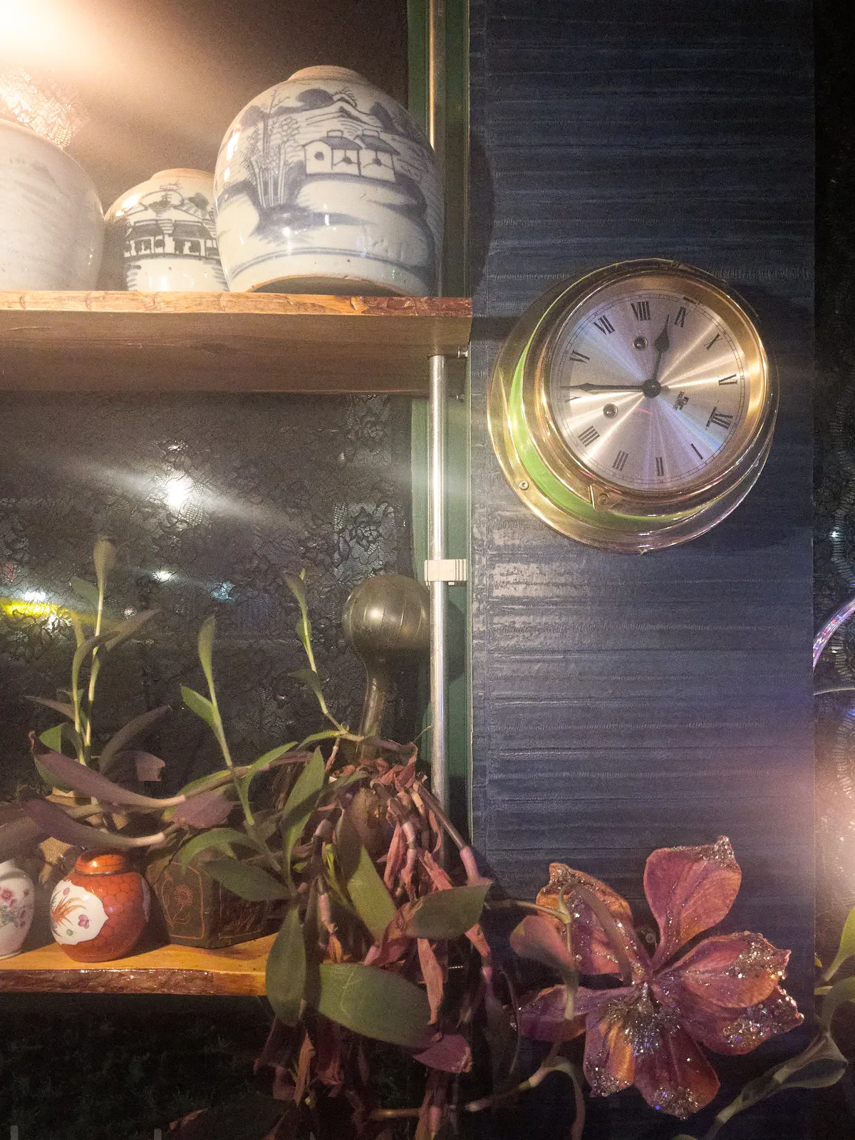 A metal wall clock is attached to a blue wall. It is surrounded by plants and ceramic pots. It points at 14 minutes to one. But when you look closely you can see that the twelve is not pointing up. So when you look closely, it is 26 minutes past 8.