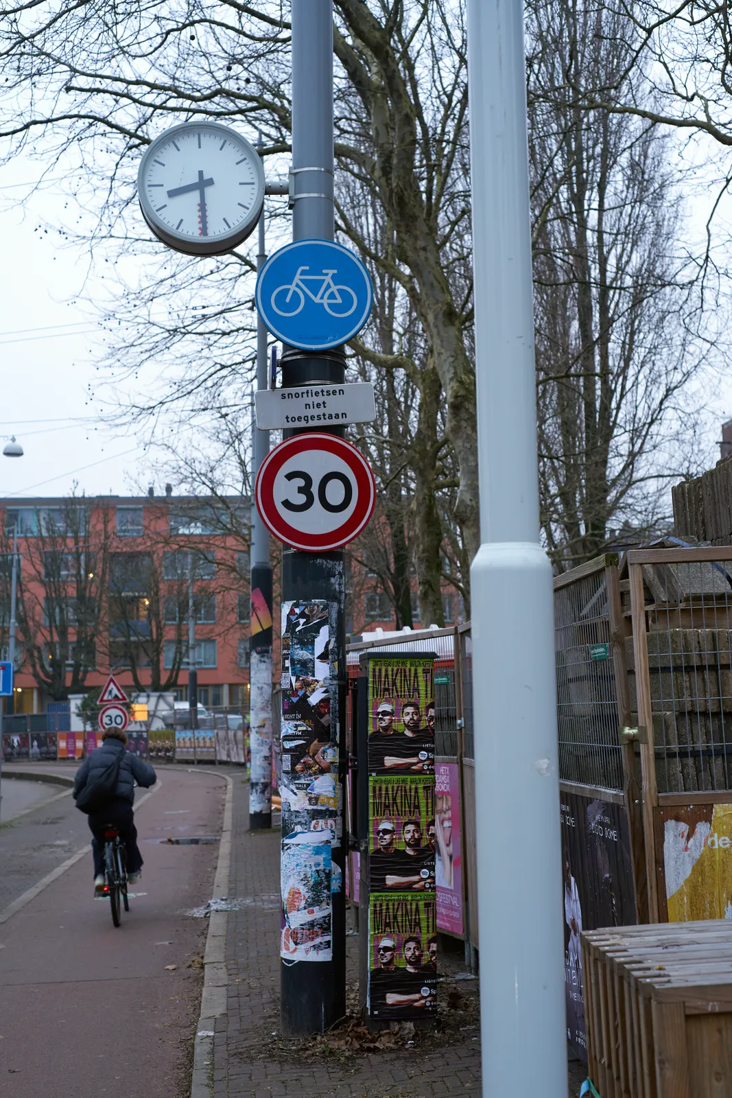 Somebody is riding their bicycle on a bike lane, seen from the back. They are riding past some fences around a work site. Next to the cyclist there’s a large pole with street signs and a clock attached to it. The clock points at 29 minutes past eight. 