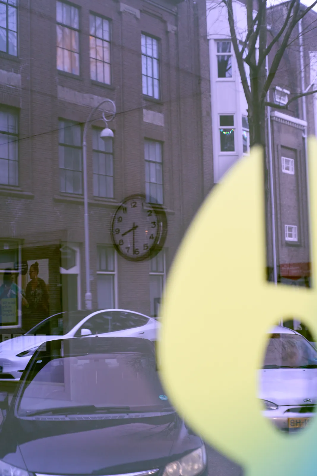 Peeking through a window. In the reflection an Amsterdam street can be seen, with cars parked in it. There’s a large yellow logo on the window, which is blurry. Inside the room there’s a clock on the wall that points at 31 minutes past eight.