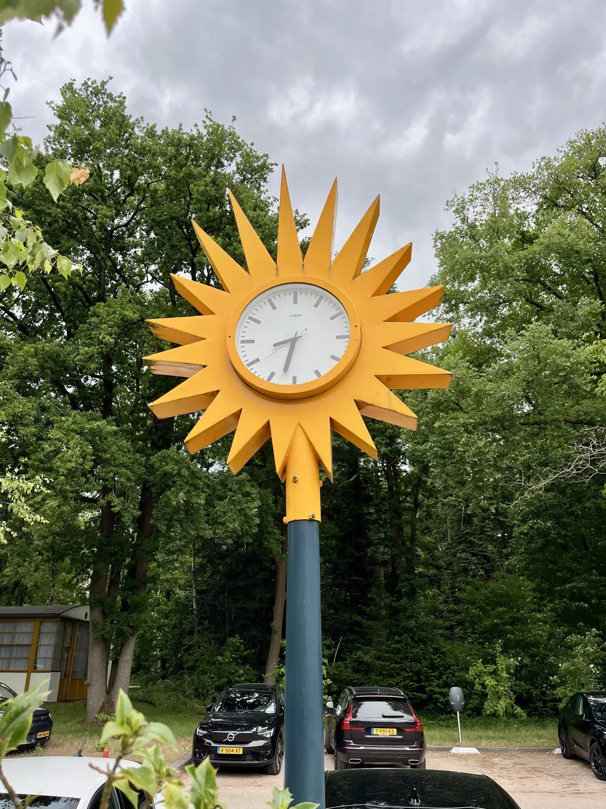 A bright yellow star (with nineteen points) with a clock in the middle, on a pole, in front of some parked cars and many, very green trees. The clock in the star points at 32 minutes past eight. 