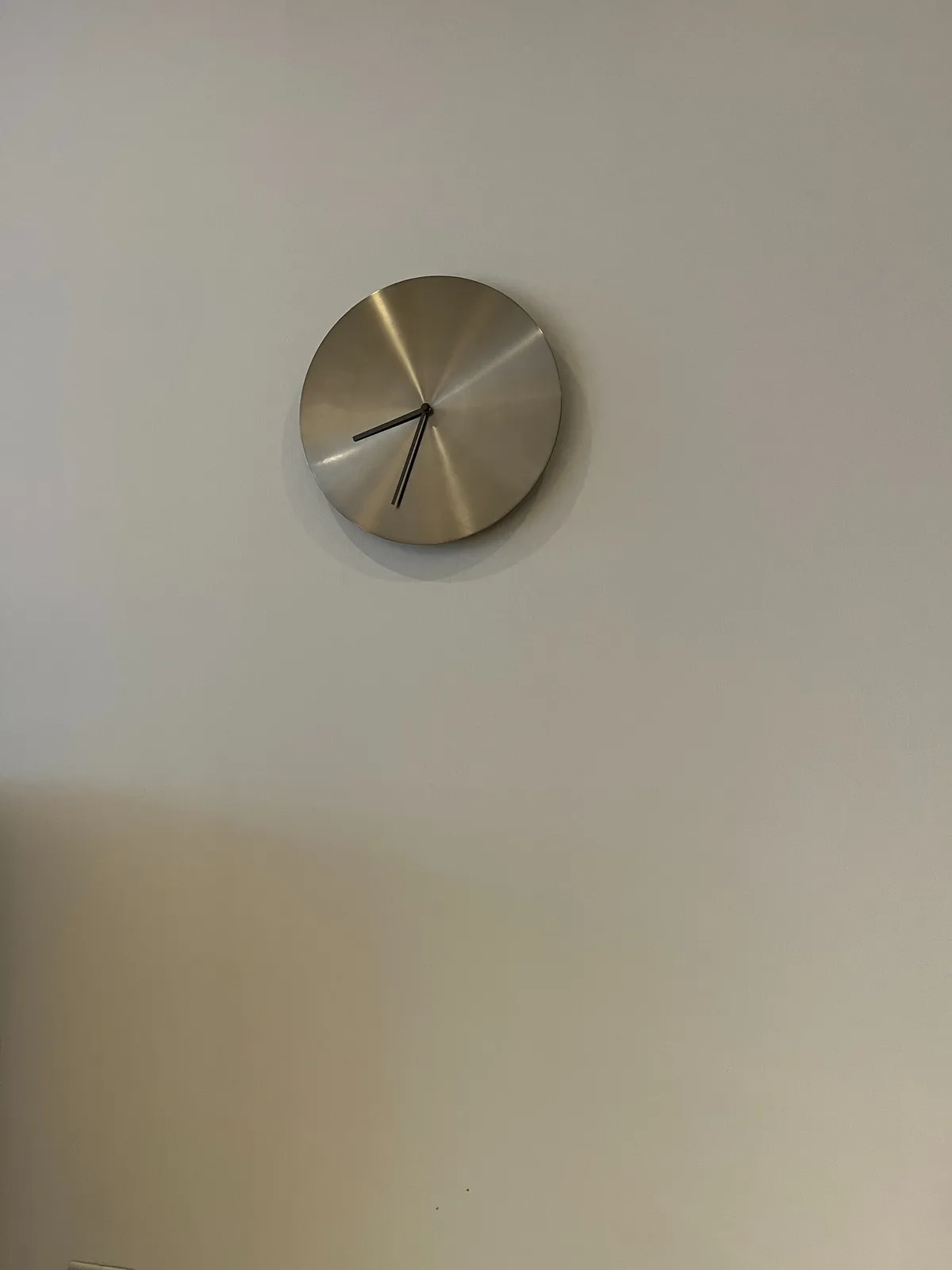 A very minimalist metal clock with black hands on a white wall. It points at approximately 33 minutes past eight.