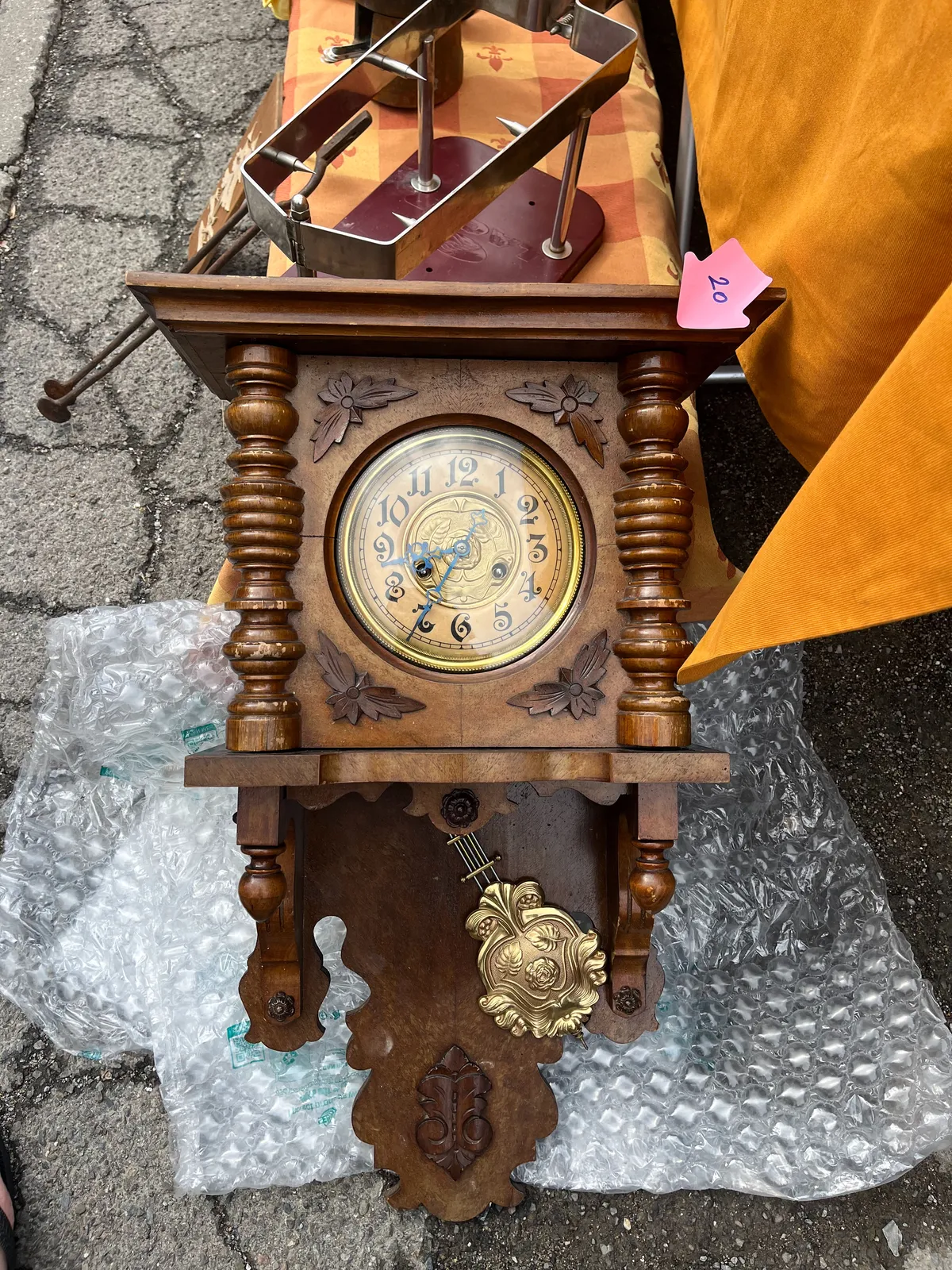An old wooden and brass clock lies on the ground. It’s for sale for 20. The wood is ornamented with some simple leaves, the brass face of the clock is ornamented with a bit more detail. It points at 25 minutes to 9.