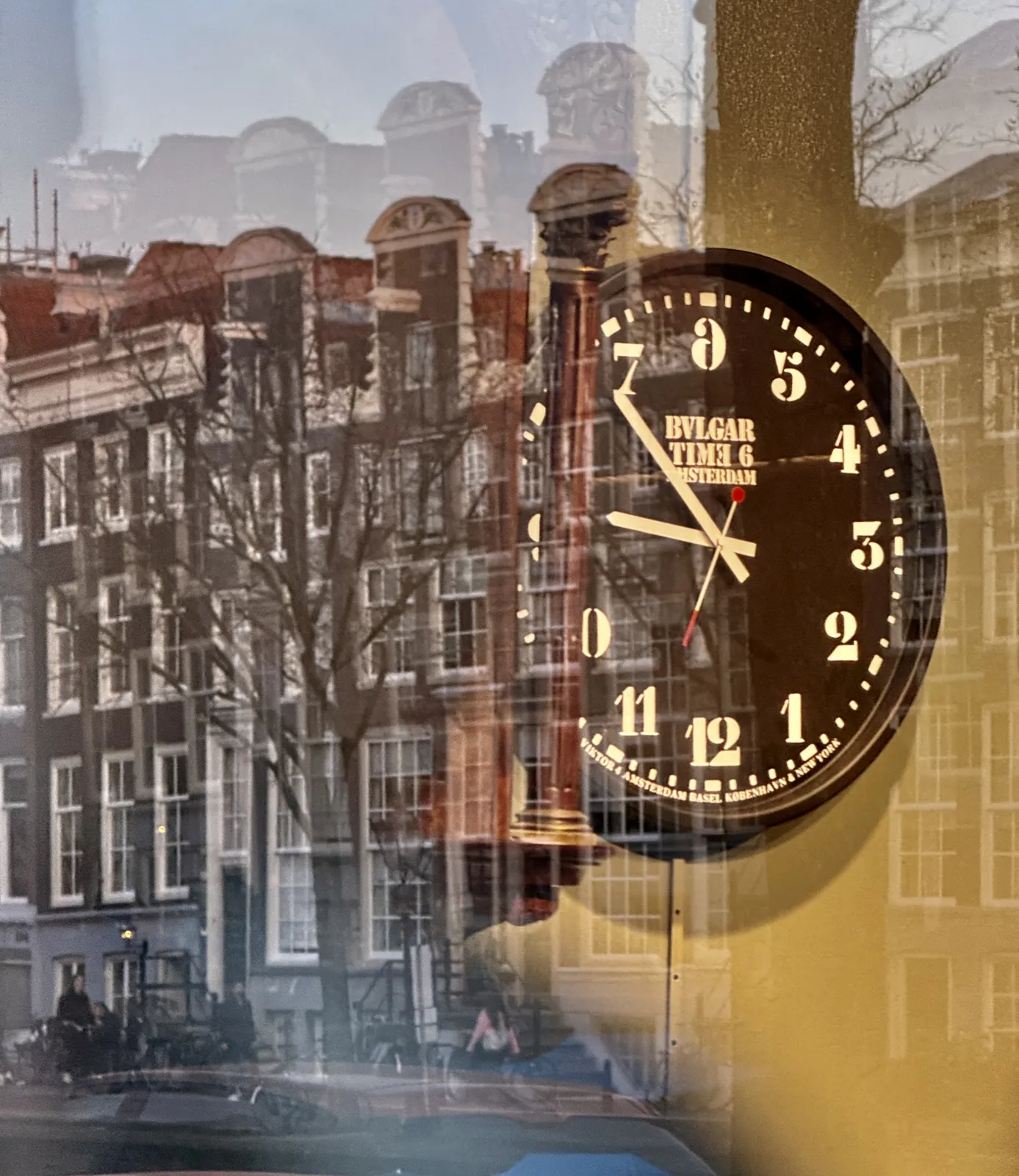 Behind a window there’s a confusing clock. In the reflection of the window there is a row of classic Amsterdam canal houses. The clock is confusing. The six is at the top, instead of the twelve, and the twelve is at the bottom. It also runs counter clockwise, which makes it extra confusing. Once you get it, you can see that it points at 36 minutes past eight.