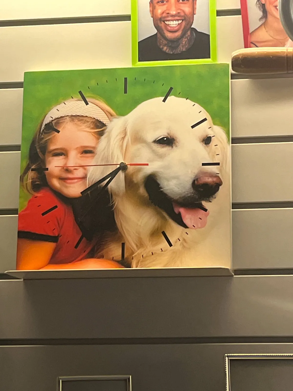 Photographic portraits of random people on a wall. One of the portraits is of a young girl with a dog. It’s not just a portrait, it’s also a clock. It points at 23 minutes to 9.