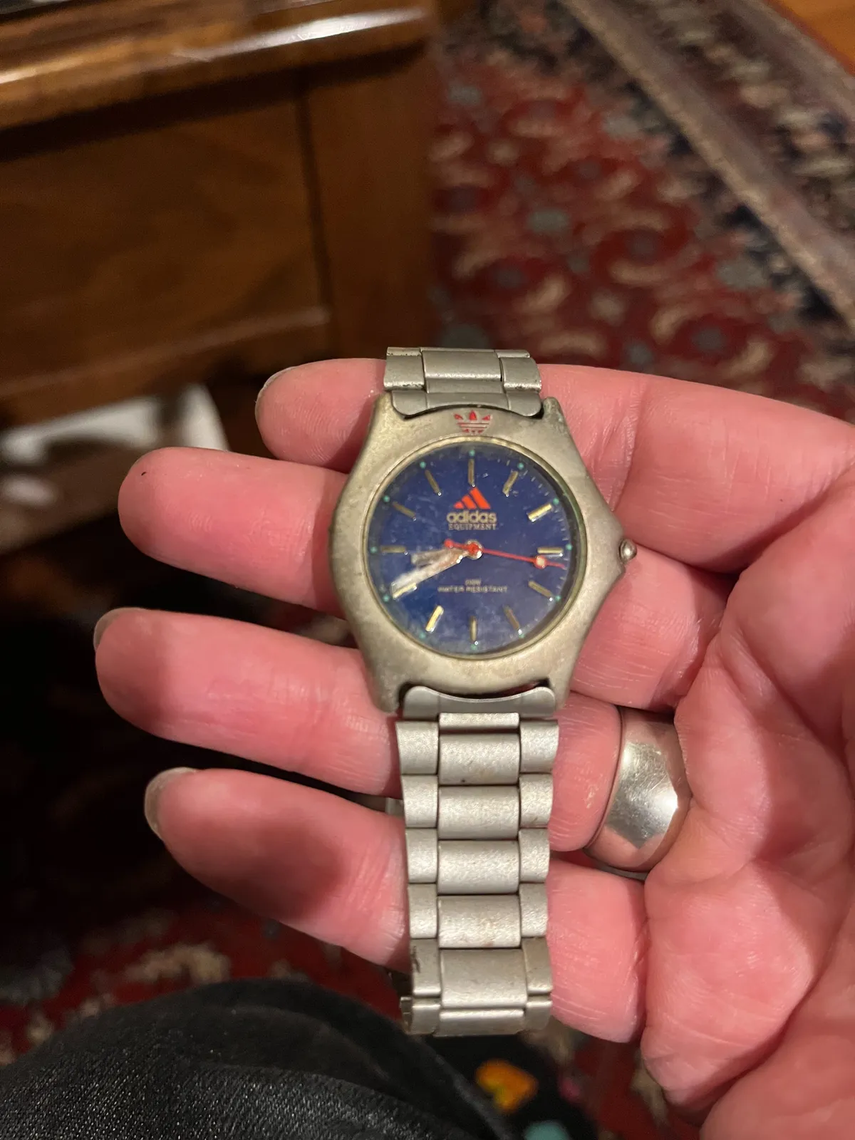 Someone is holding an old watch in their hand. In the background, blurry, an old Persian rug. The watch points at 20 to nine.