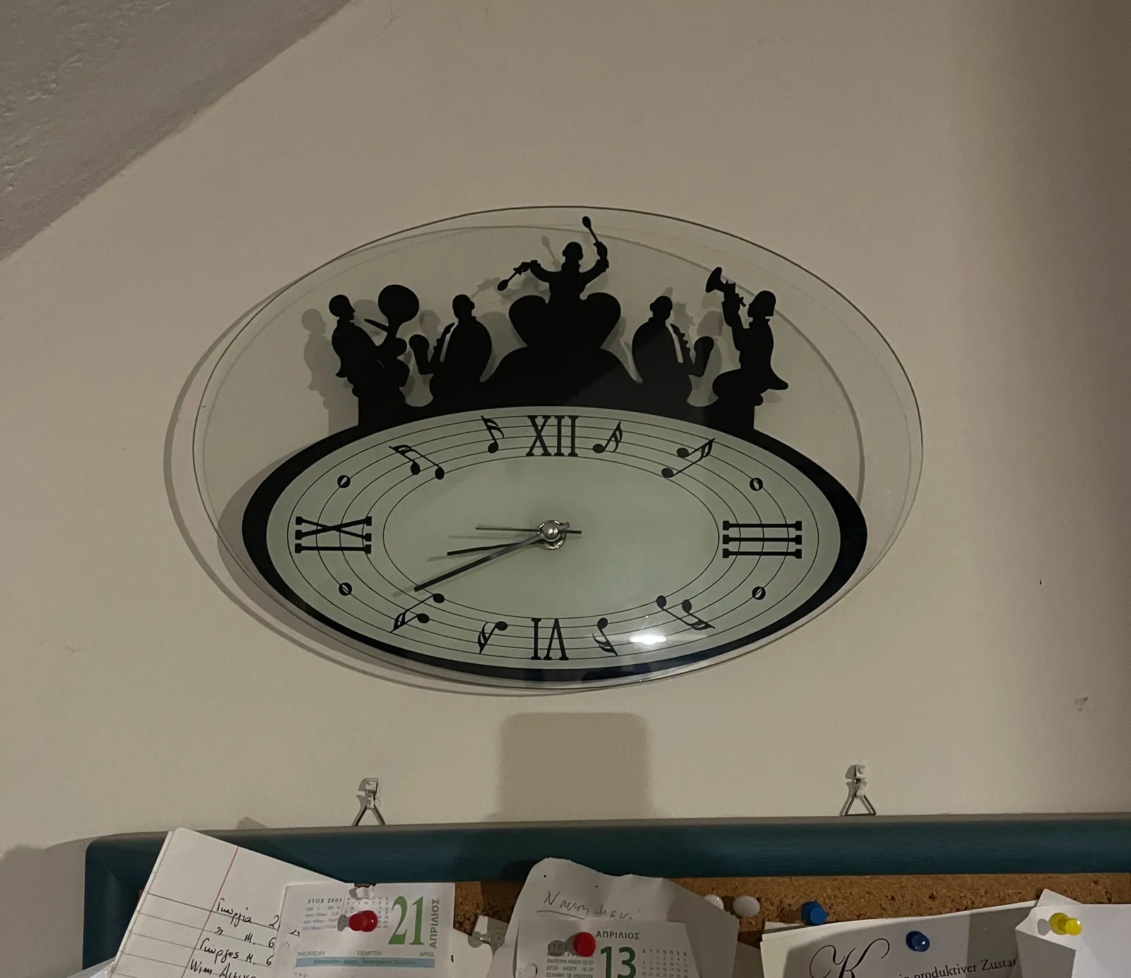An oval shaped clock with roman numerals that indicate three, six, nine and twelve. Musical notes indicate the other numbers. It points at approximately 19 minutes to nine.