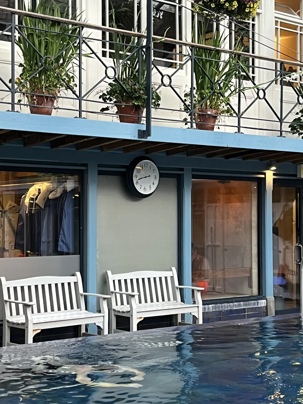 Behind a swimming pool there are two white, wooden benches, and a wall clock attached to the wall behind them. This clock points at 18 minutes to nine.