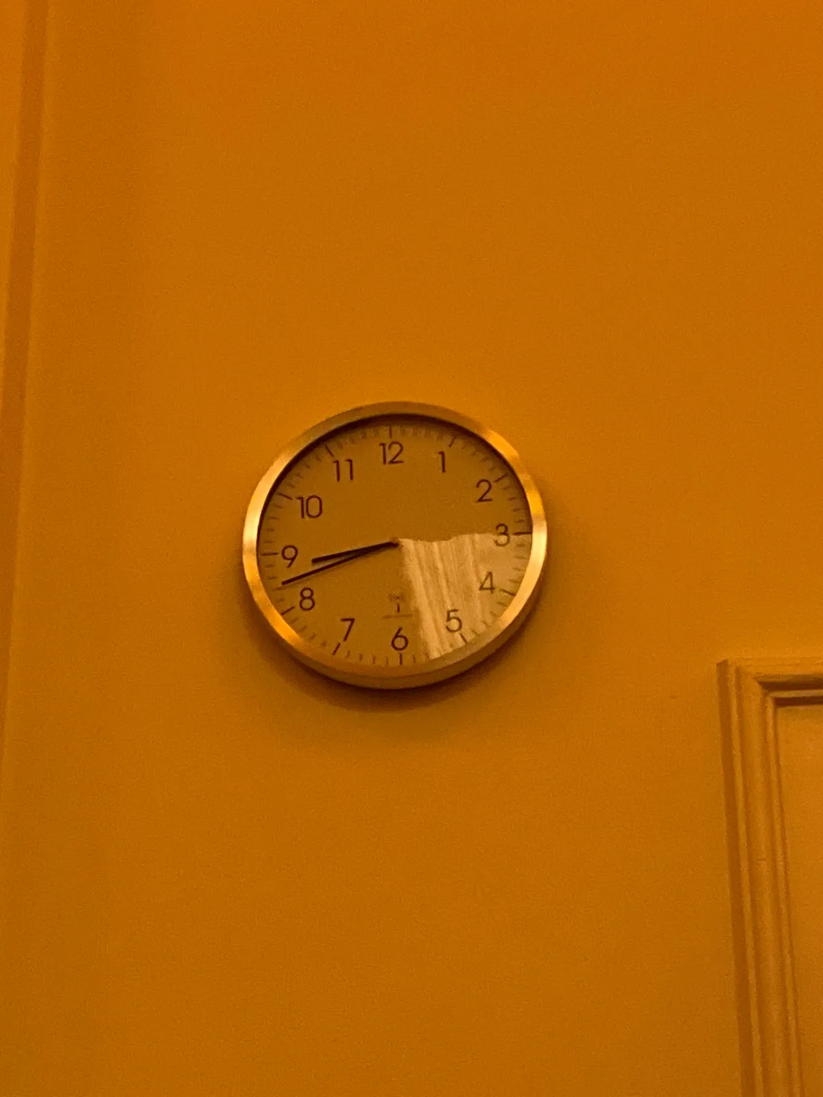 A corner of a door can be seen in a corner of this image. The rest of the image is a yellow wall with a clock in the center. On this clock it is 18 minutes to 9.