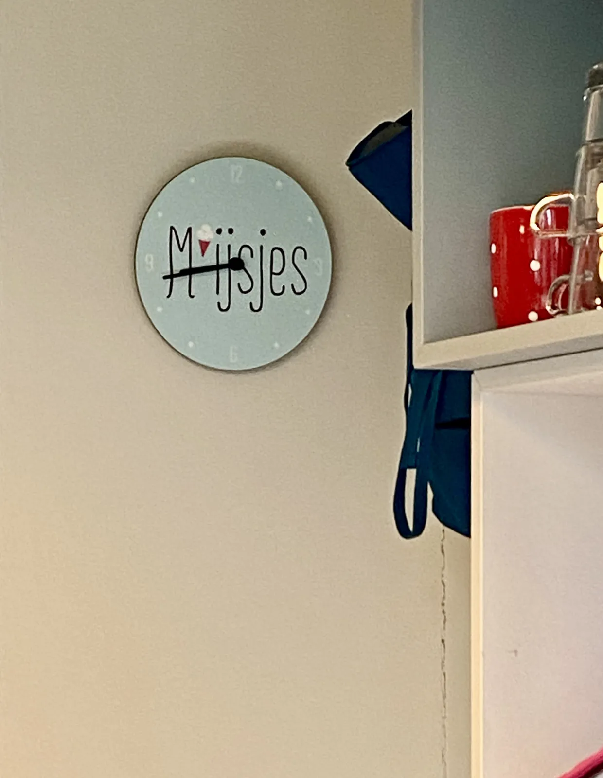 A clock, next to a cupboard with colourful mugs in it. On the clock it says M ijsjes. This is a dutch play of words. It means, phonetically, on the one hand, something like hm, nice, ice cream! Read differently, it means Girls. This clock points at 17 minutes to 9.