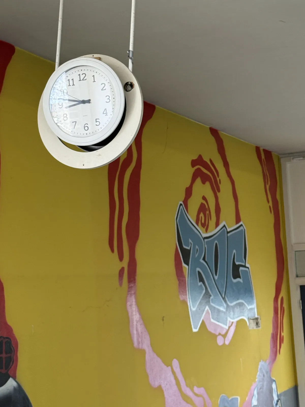 Someone painted a gaffiti piece on the wall behind this clock that hangs from the ceiling. It points at 14 minutes to nine.