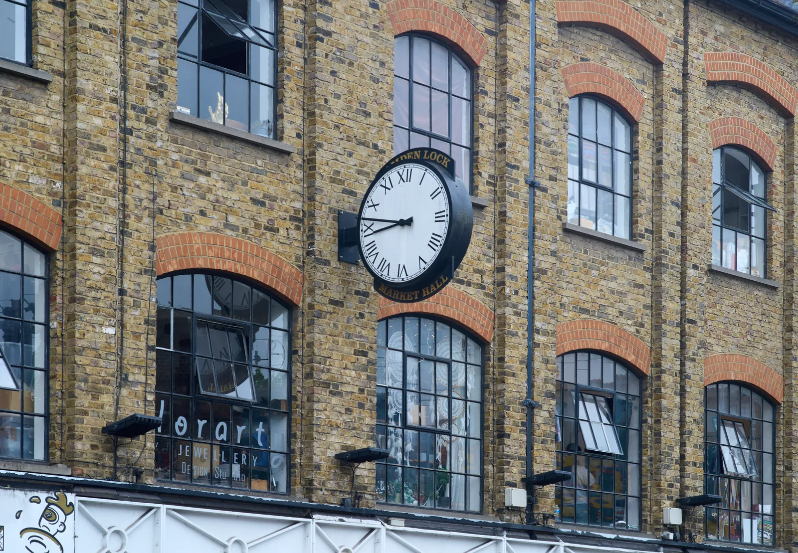 Attached to the facade of an old industrial brick building with high windows, is a large black clock. The words below it say Market Hall, only the word Lock can be read from the words above it. The clock points at 13 minutes to nine.