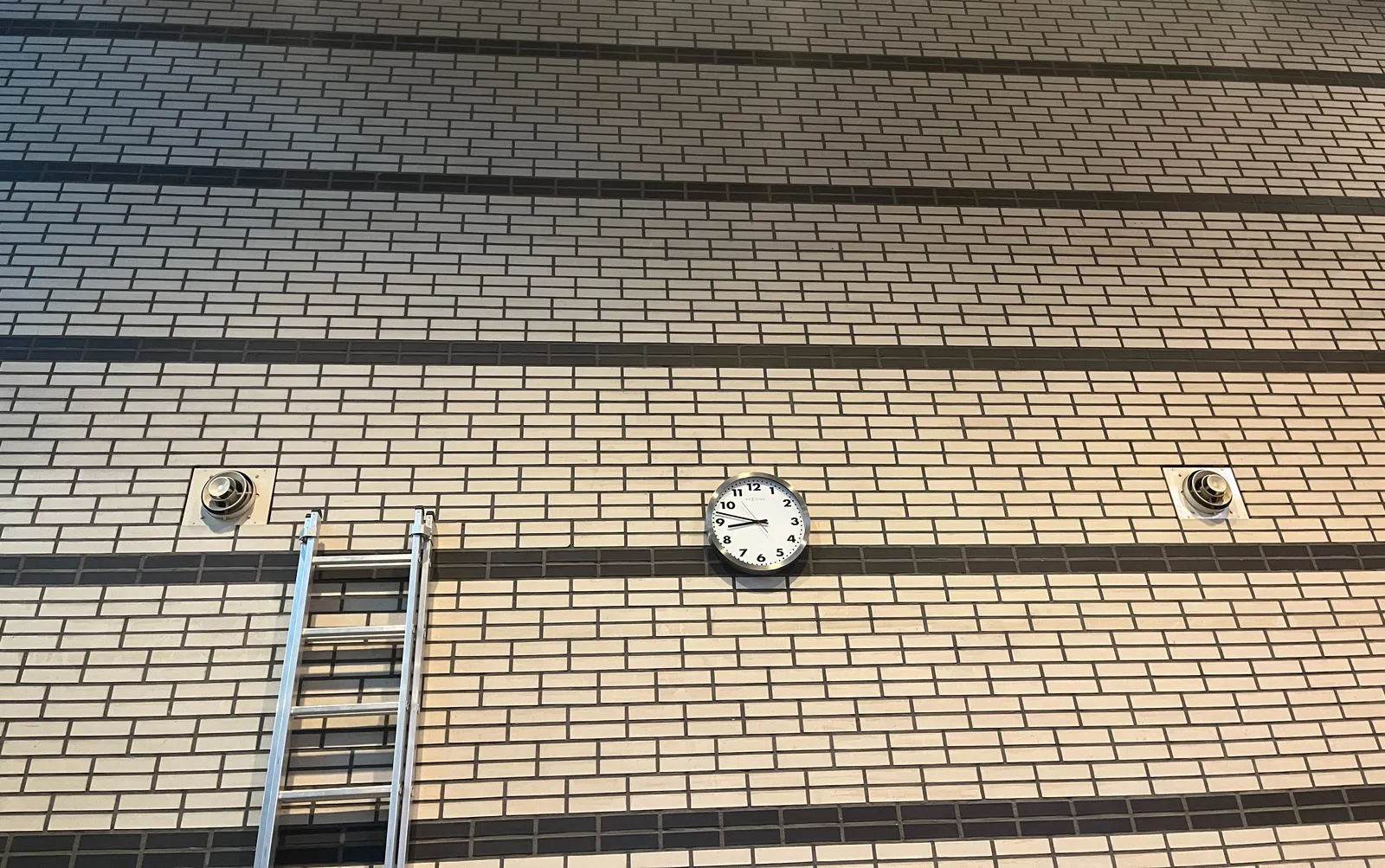 A wide and high, white brick wall with stripes made from black bricks. There’s a ladder on the left, and a clock in the middle. It points at 13 minutes to 9. 