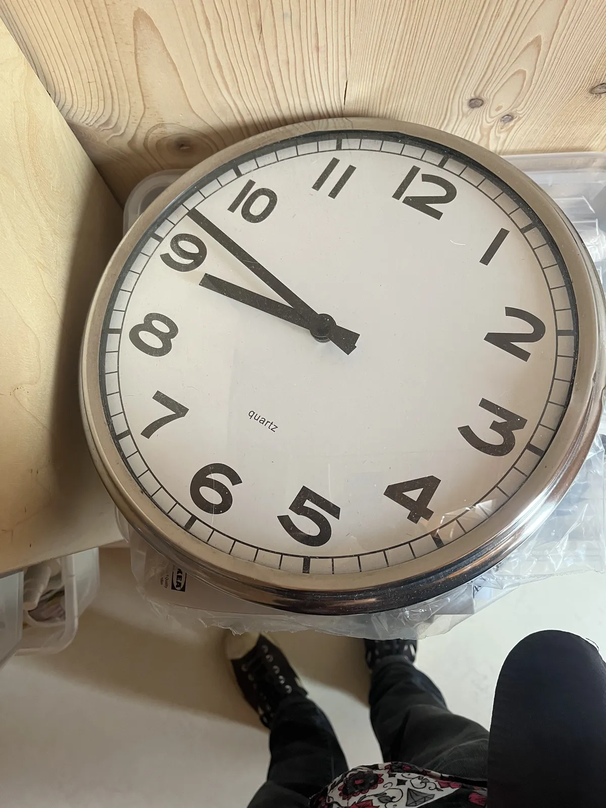 A large modernist wall clock, laying on a shelf. Below the shelf the feet of the photographer can be seen. The clock points at 13 minutes to nine.
