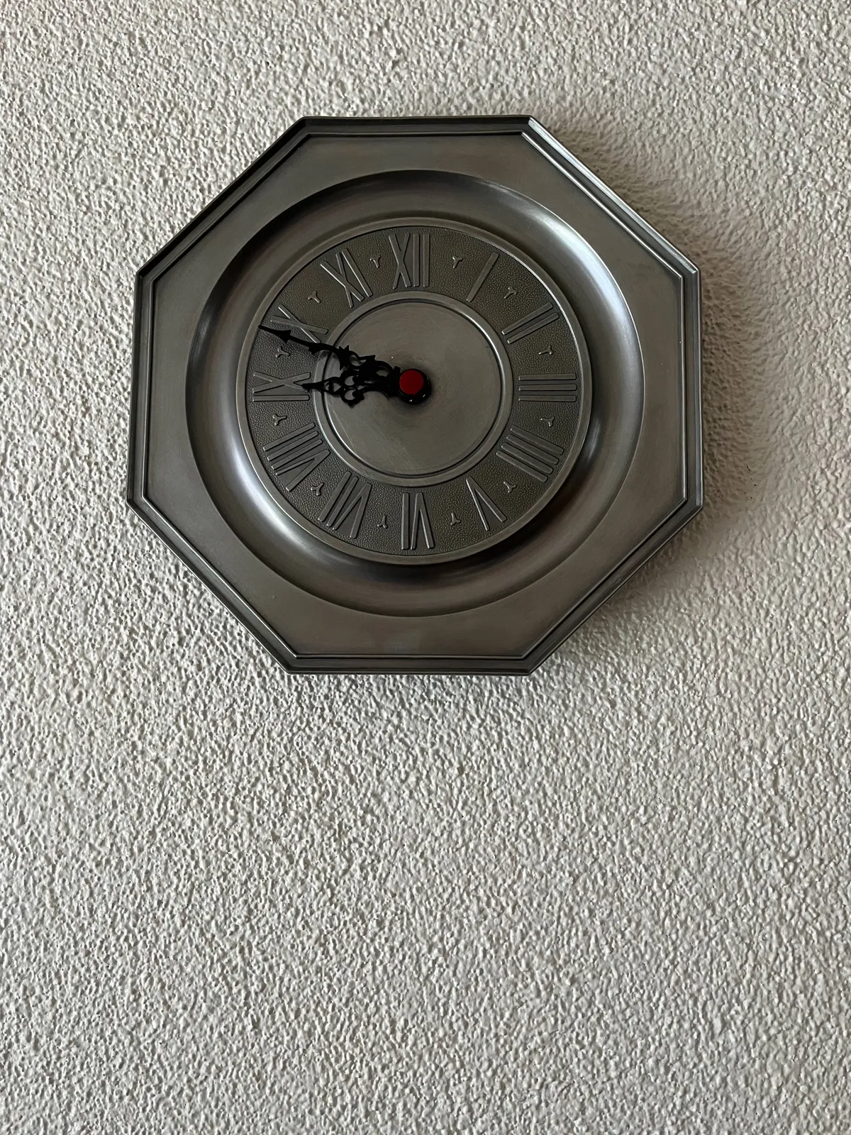 A metal wall clock, silver coloured, with the timestamps embossed in roman numerals. The clock points at 11 minutes to 9