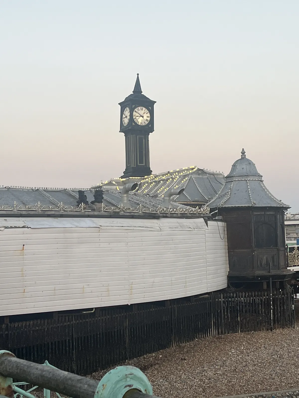 A clock in the shape of the Big Ben, but much smaller, stands on some wooden shed-like structures with party lights on them. The clock on this small big ben points at 9 minutes to nine.