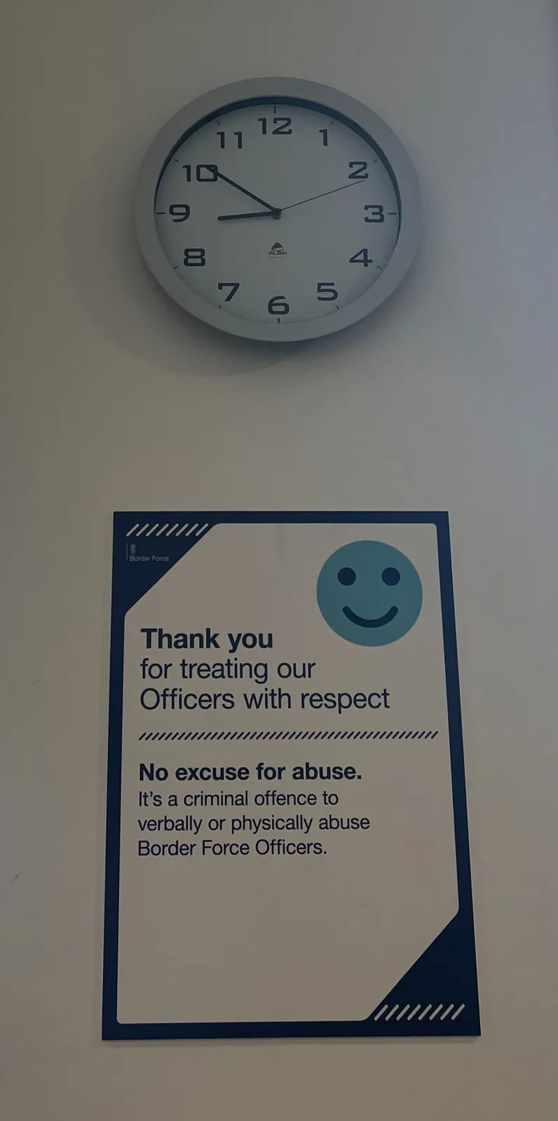 There’s  a sign on the wall that says: Thank you for treating our Officers with respect. No excuse for abuse. It's a criminal offence to verbally or physically abuse Border Force Officers. Above this sign there’s a clock that points at nine minutes to 9.