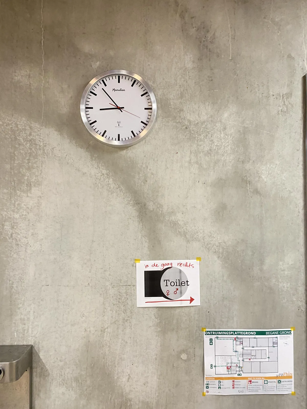 A concrete wall with a safety escape plan, a sign showing where the toilet can be found and a clock. The clock points at 7 minutes to 9.