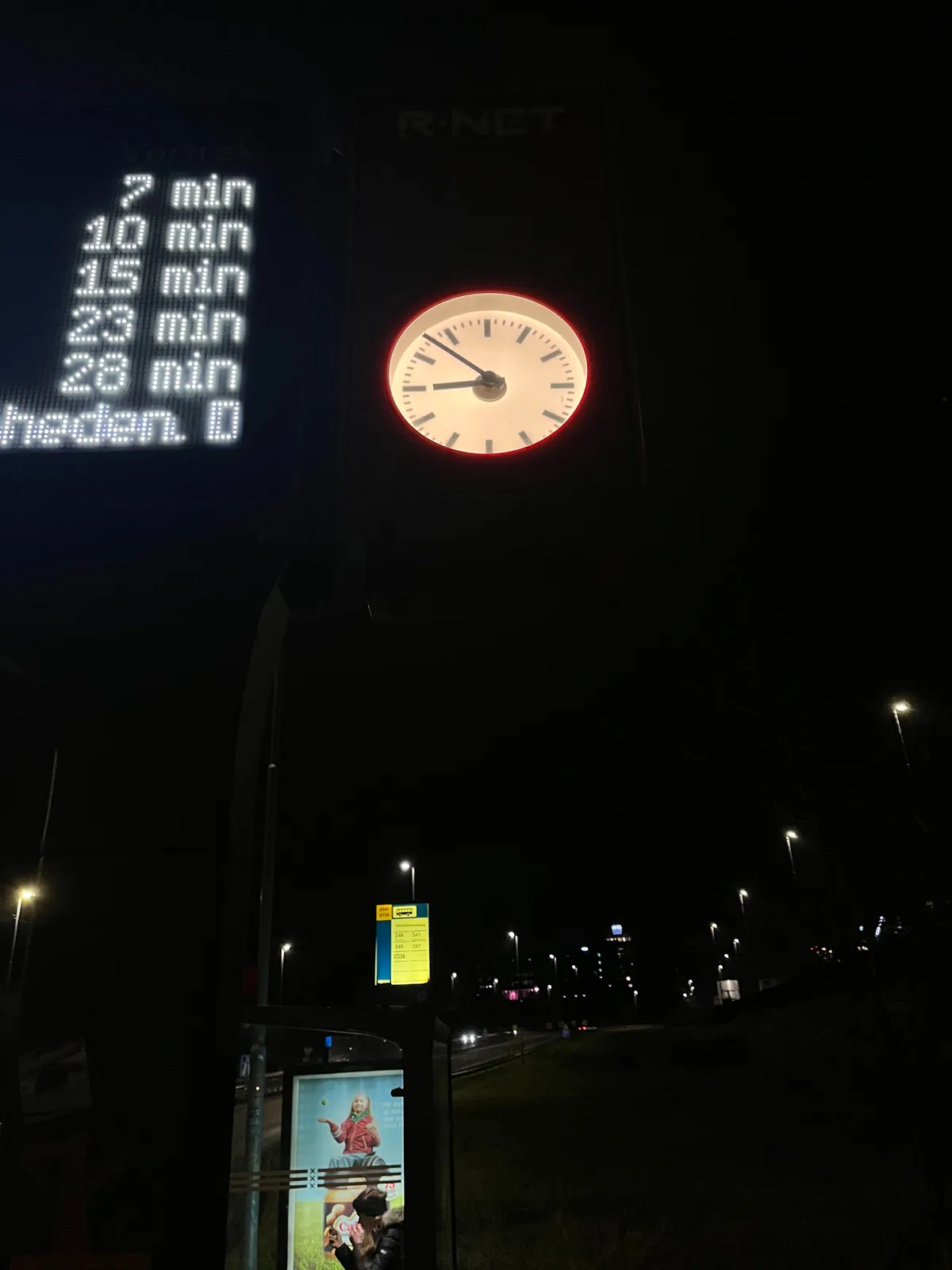 It’s dark. In the background some street lights and a bus stop. In the foreground, at the top of the picture there’s a clock that points at 7 minutes to 9. There’s also part of a display. It says 7 min, 10 min, 15 min, 23 min and 28 min. Probably how long it takes before busses arrive.