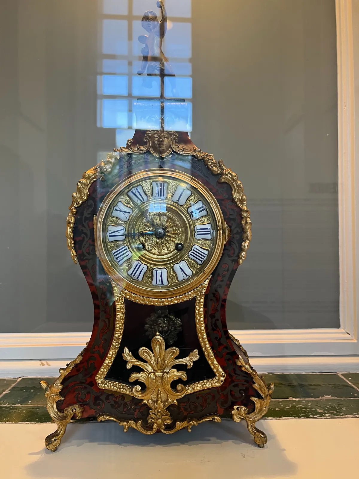 A jubilant cherube stands on top of this richly ornamented mantel clock, made from burl wood and gold. The clock points at 6 minutes to 9.