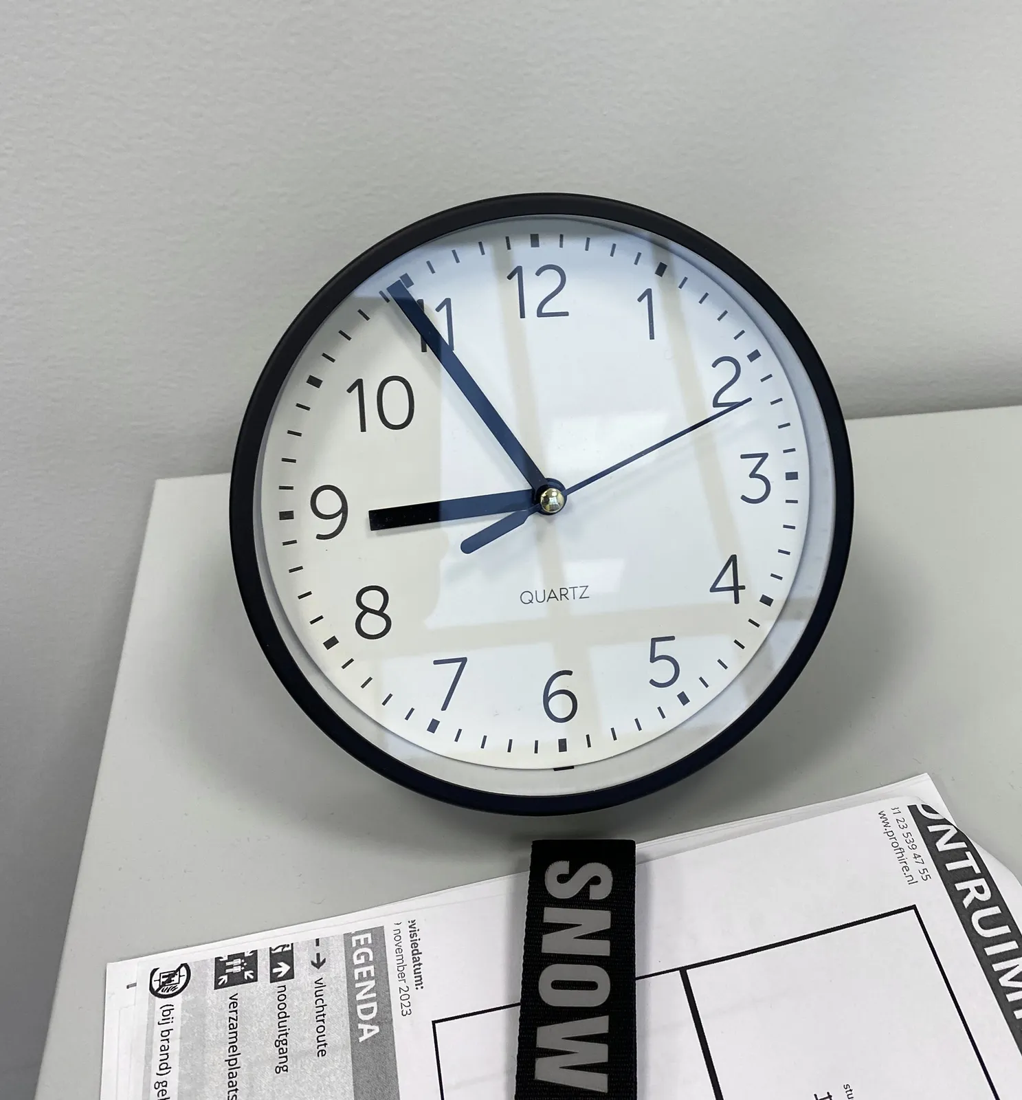 A wall clock standing on a piece of paper with stuff printed on it. The clock is black and white, the print is black and white. The clock points at 6 minutes to 9.