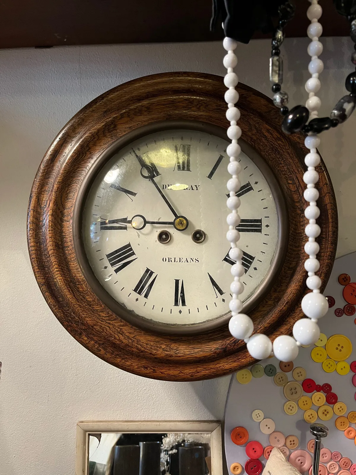 A white plastic necklace hangs in front of a clock. On the clock the time is 6 minutes to nine.
