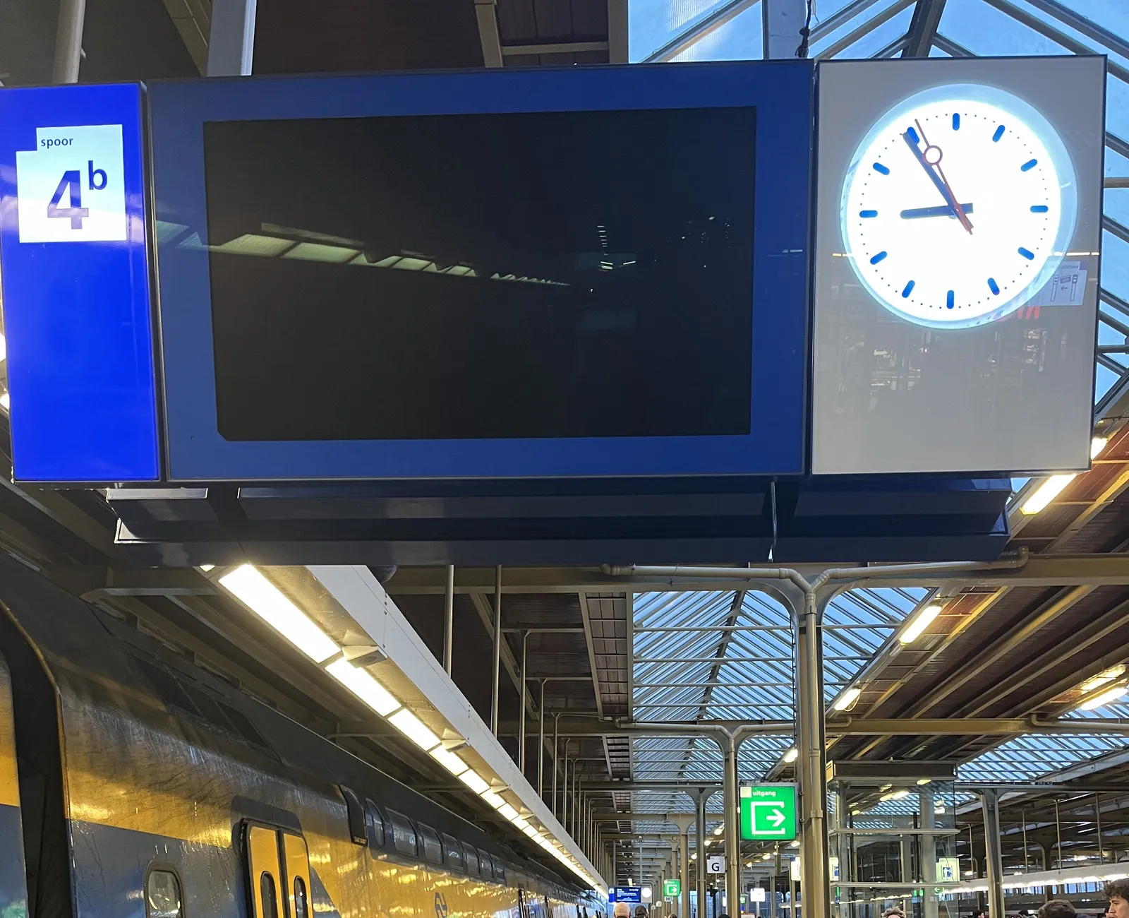 A display on a train station. Usually these displays show information about the train, this display is black. The train stands next to it, to the left. To the right there’s a station clock that points at 6 minutes to nine.
