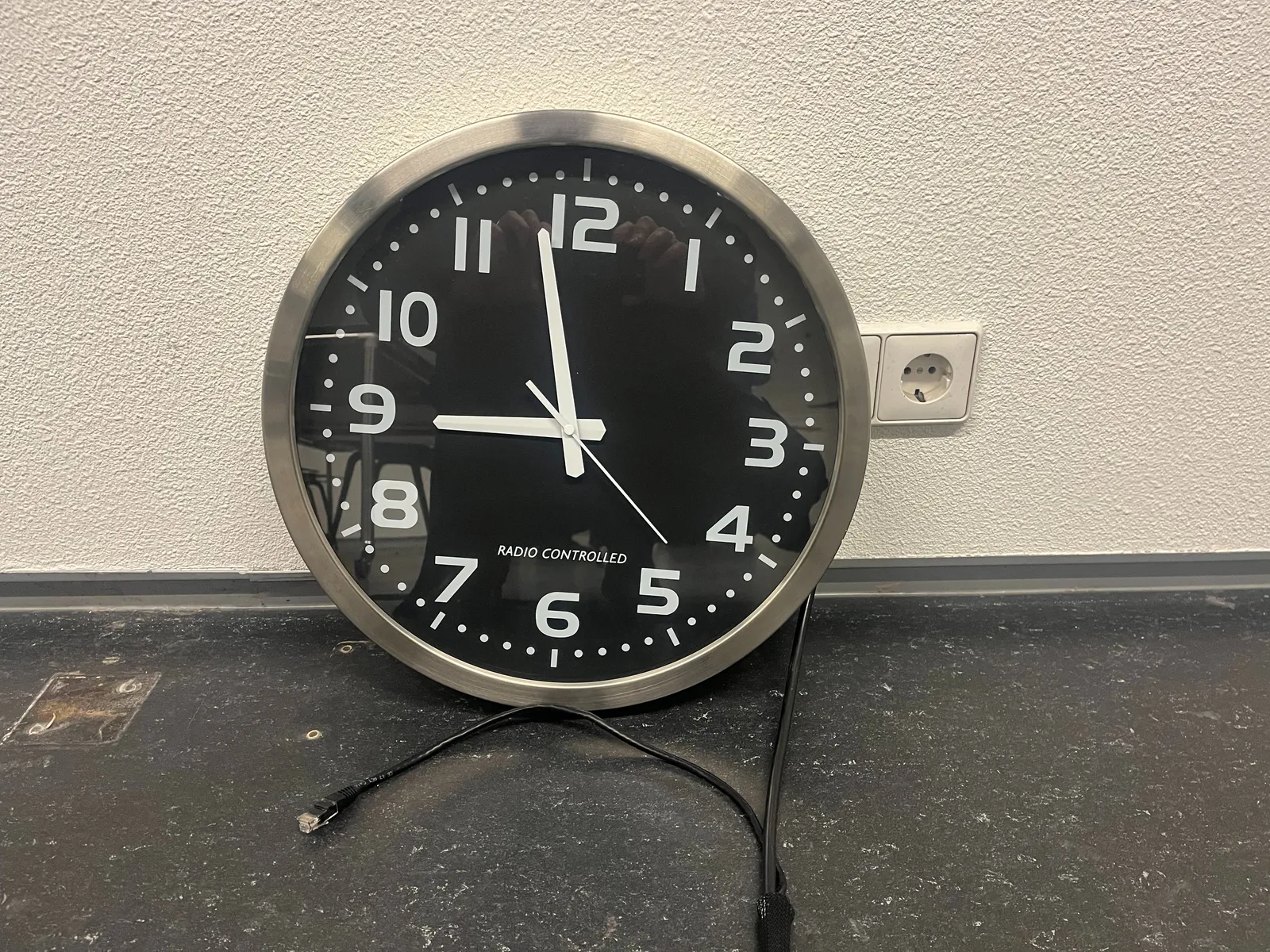 A black wall clock, standing on a black floor, leaning against a wall, next to a wall socket. The clock is radio controlled, it says on the face. It points at 2 minutes to 9.