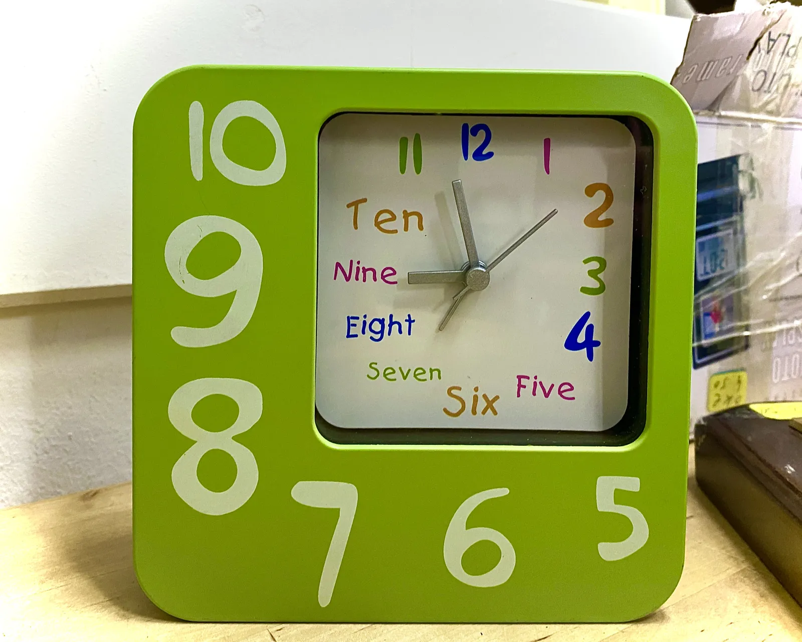 An eclectic, colourful clock. All hours are written as numbers, and some of them are also written as words. The frame is asymmetric and it’s green. The clock points at 2 minutes to nine.