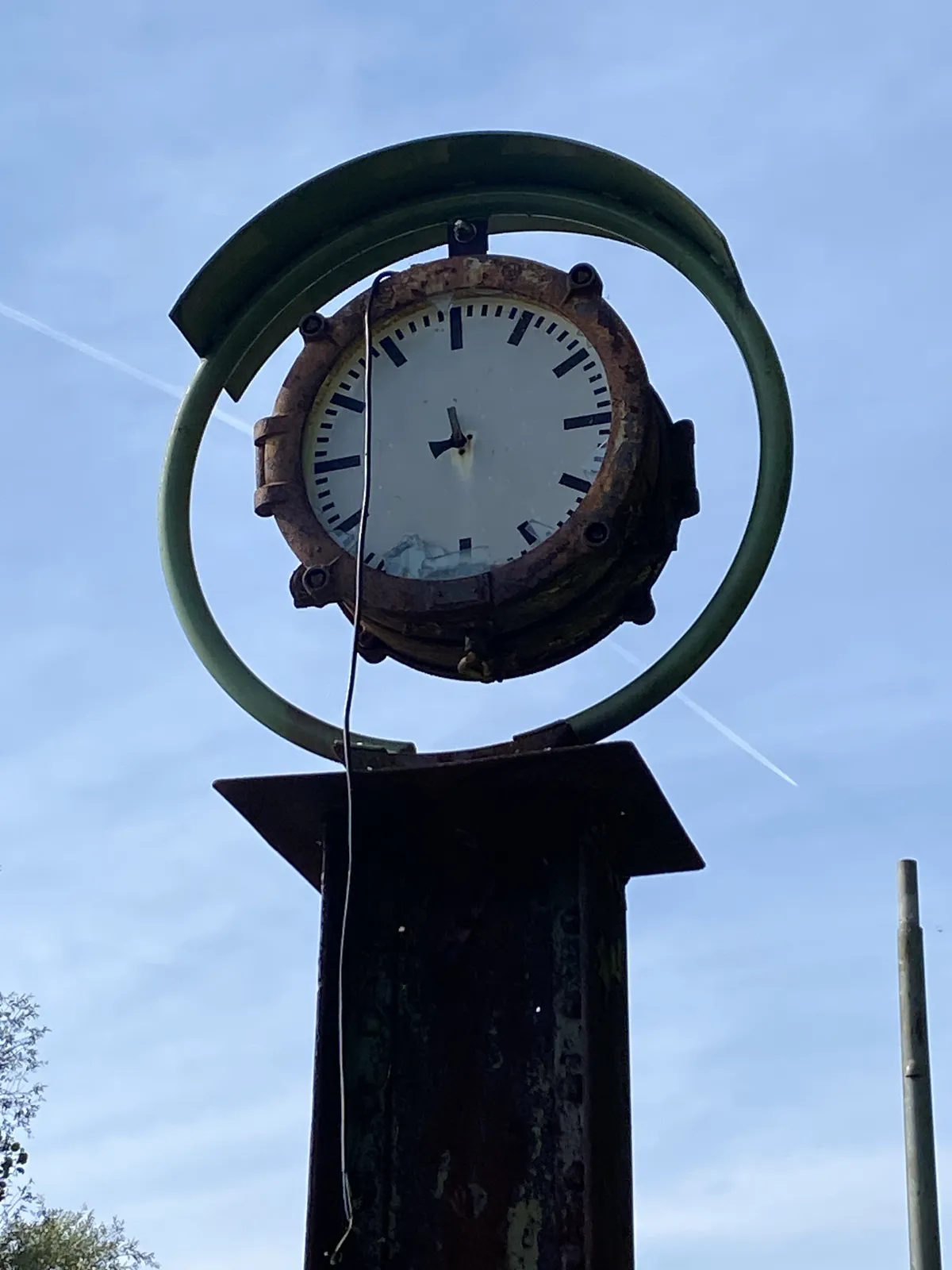 A clock in a green round frame, mounted on a simple piece of construction metal. The hands of the clock are broken, so it’s hard to tell what time it is exactly. It seems to be pointing at 2 minutes to 9.