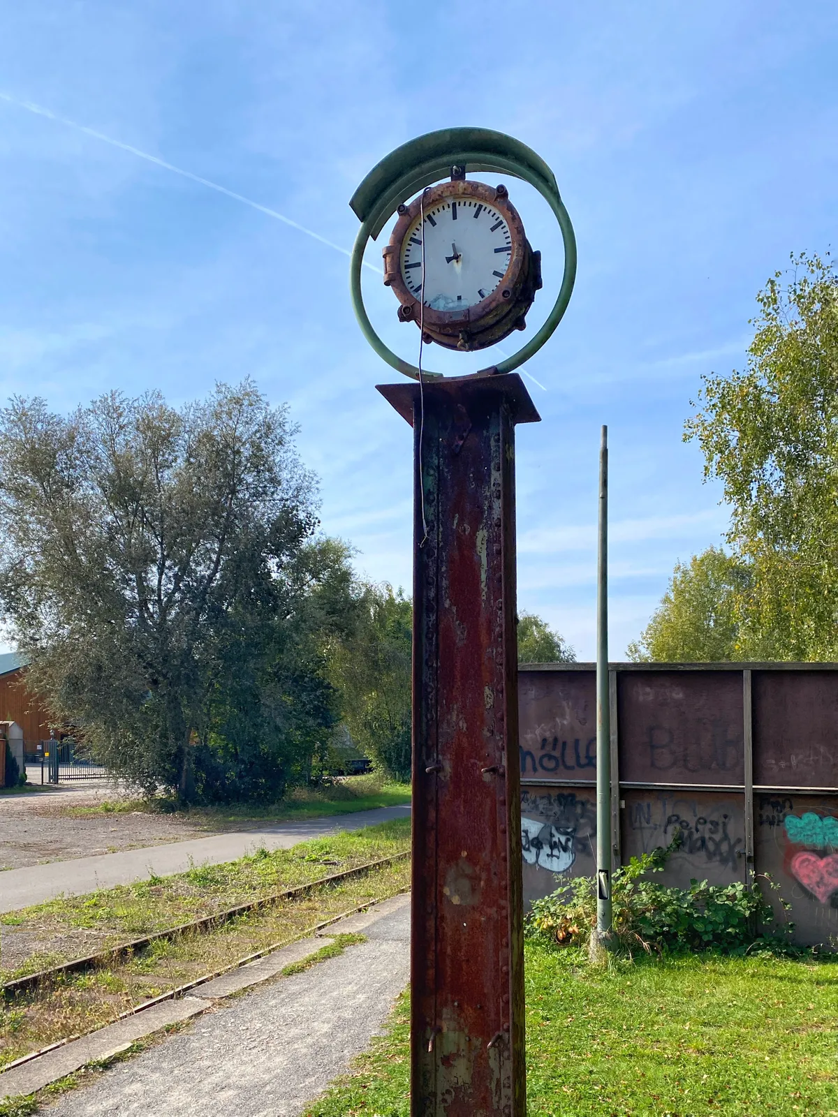 A clock next to a train track. The clock is mounted on a simple piece of construction metal. The hands of the clock are broken, so it’s hard to tell what time it is exactly. It seems to be pointing at 2 minutes to 9.