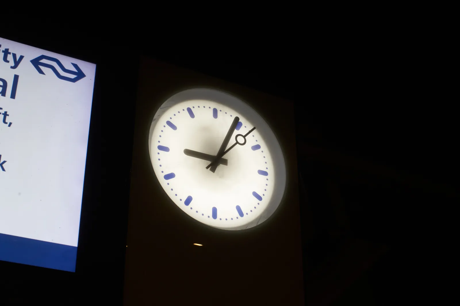 A clock in a train station. The clock is brightly lit, around it it’s dark. The clock points at 4 past 9. 