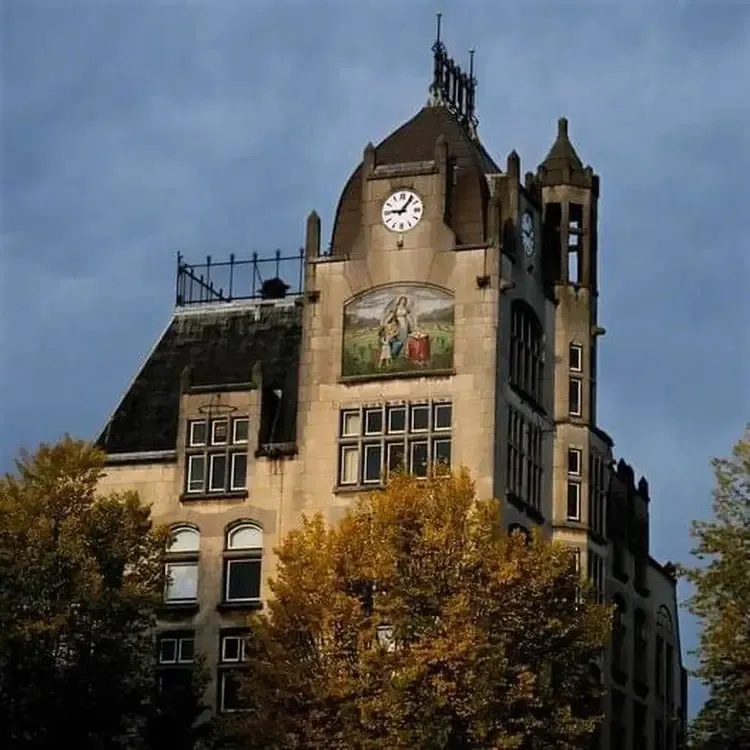 A large asymmetric building, behind some trees. There’s a painting on this building of a kid and a grown up, or maybe an angel, standing in a garden.. There’s also a clock on this building which points at 5 minutes past 9.