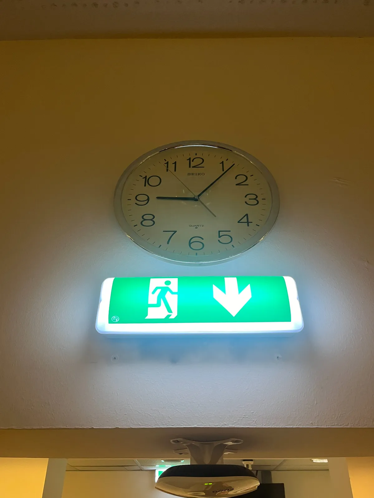 A clock right above a brightly lit exit sign. It points at 6 minutes past 9.