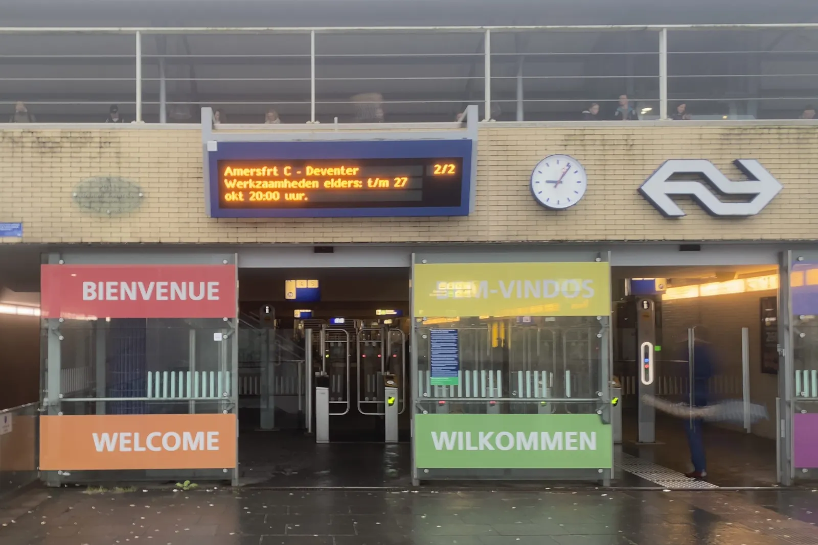 A trainstation with large coloured signes that say welcome in at least four different languages. There’s also a sign that tells about disruptions. And there’s a clock that points at 6 minutes past 9.