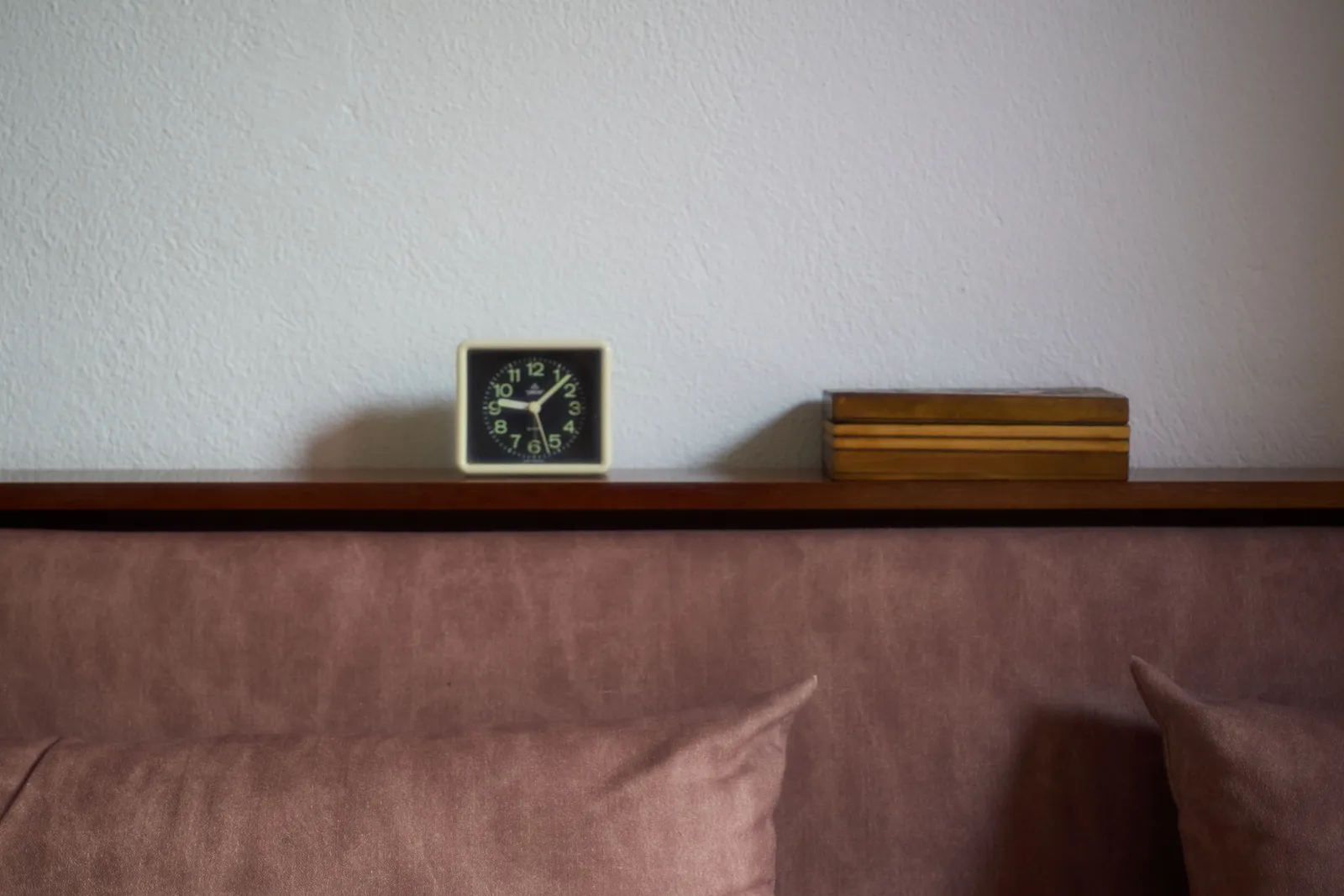 On a couch stands a wooden box, next to a small alarm clock that points at 7 minutes past nine.