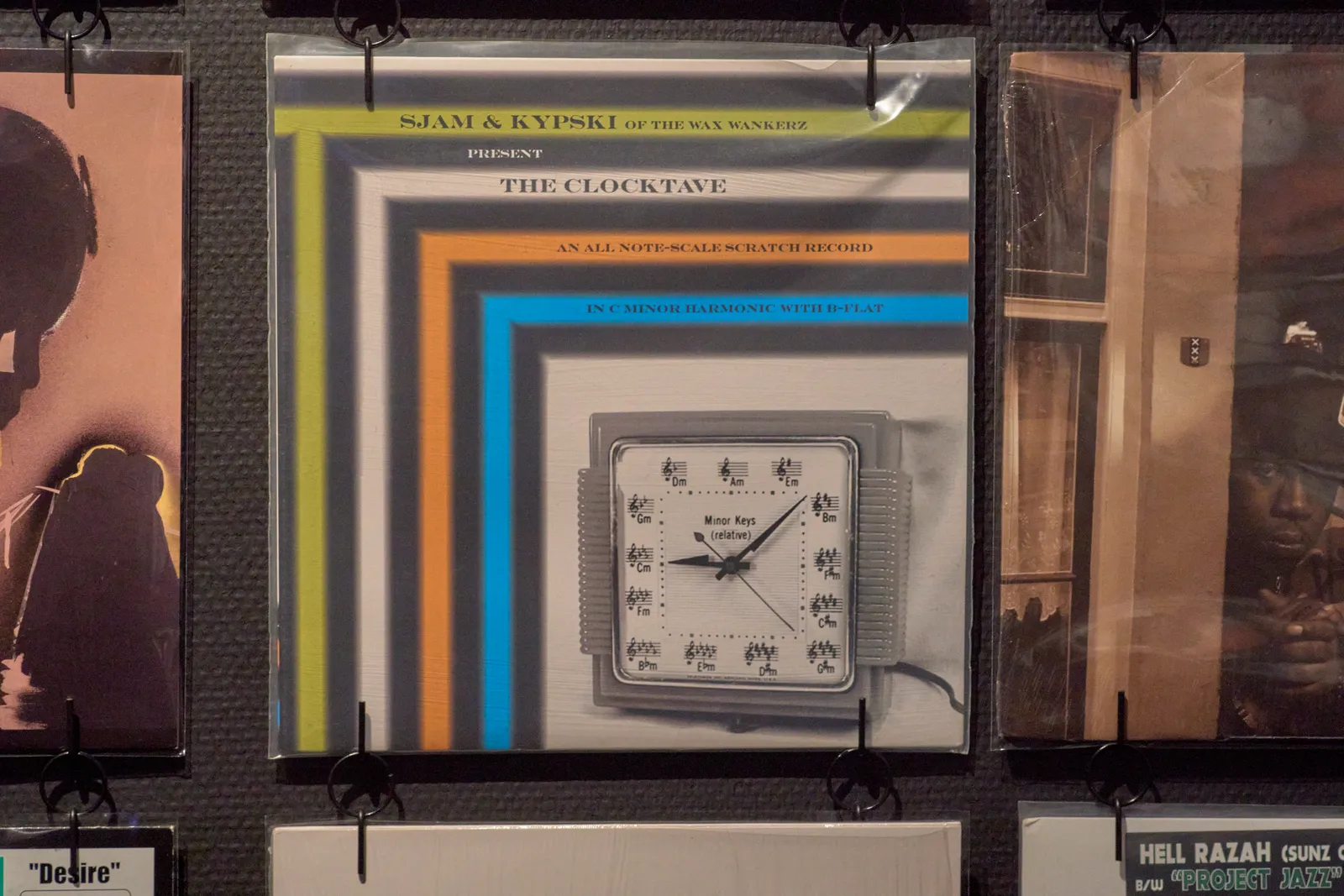 The cover of a vinyl record on a wall, among other covers. There’s a colourful pattern on it, and it’s titled Sjam & Kipsky of the wax wankerz present the clocktave, an all note-scale scratch record, in C-minor harmonic with b-flat. There’s also a clock with musical notes instead of numbers. This clock points at 8 minutes past nine.