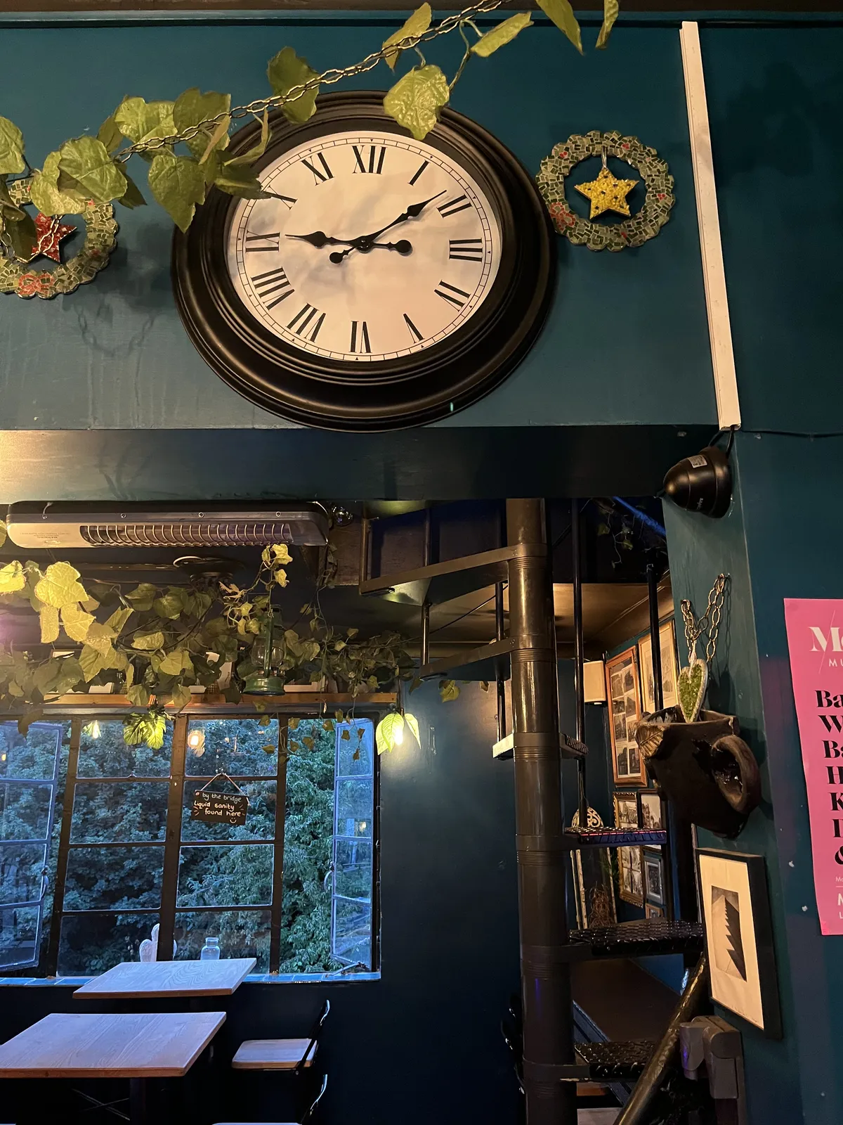 A clock above a door in a teal room, with some plants. Outside, through the window, a green tree can be seen. The clock points at eight minutes past nine.