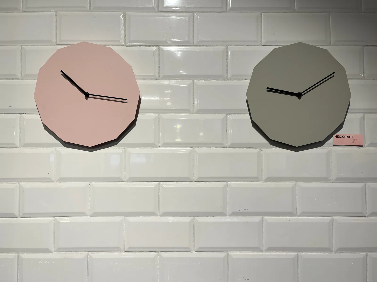 There are two clocks, both with twelve corners, hanging on a wall made from shiny white tiles. The pink clock, on the left, points at approximately 17 minutes past 10. The other one, which is green, points at 9 minutes past 9.