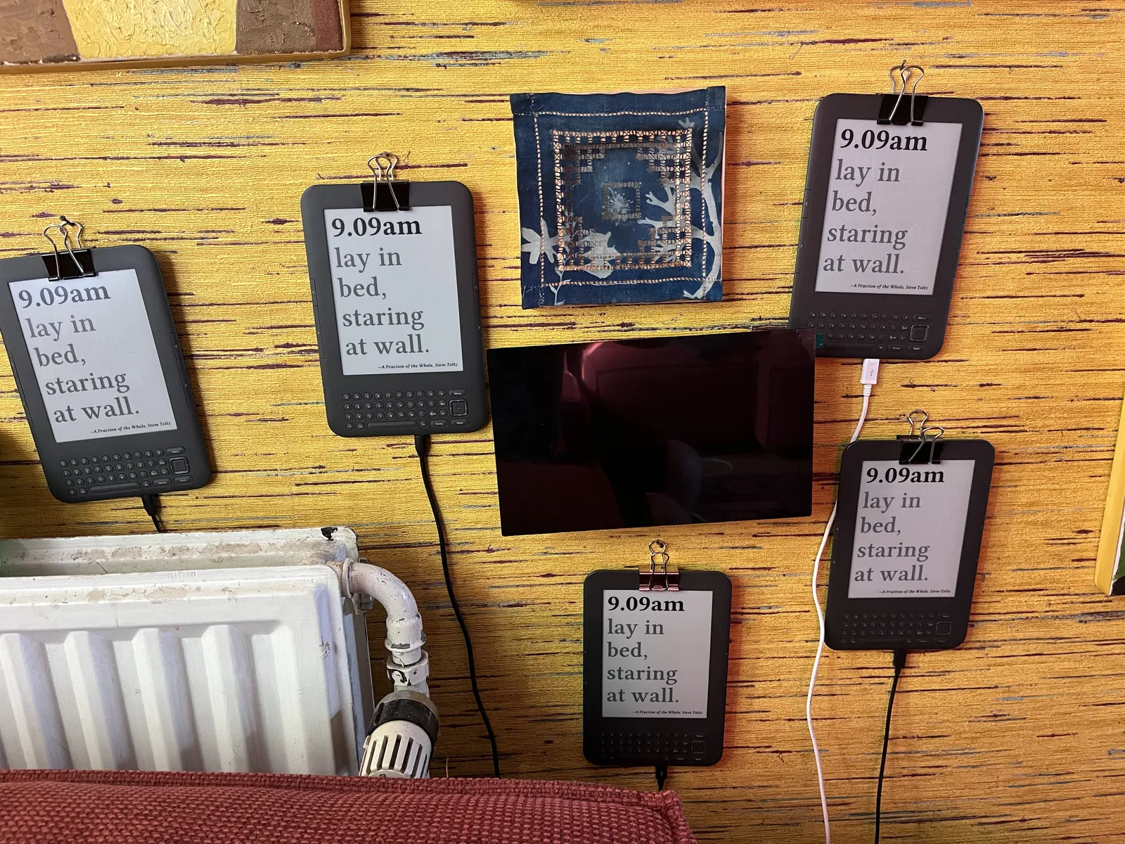 Five old e-readers hanging on a wall. On all these e-readers the same quote is printed: 9:09AM lay in bed, staring at wall.